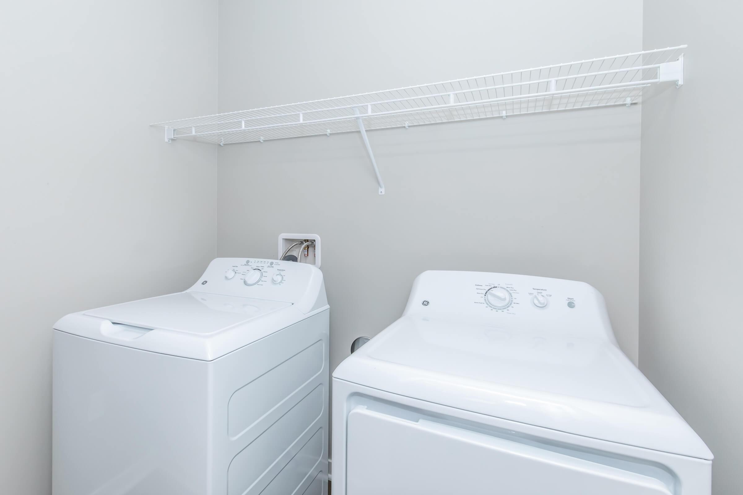 A clean laundry room featuring a white washing machine and a white dryer side by side. Above them is a metal wire shelf for storage, set against a light gray wall. The space is well-organized and bright, suggesting a functional area for household chores.