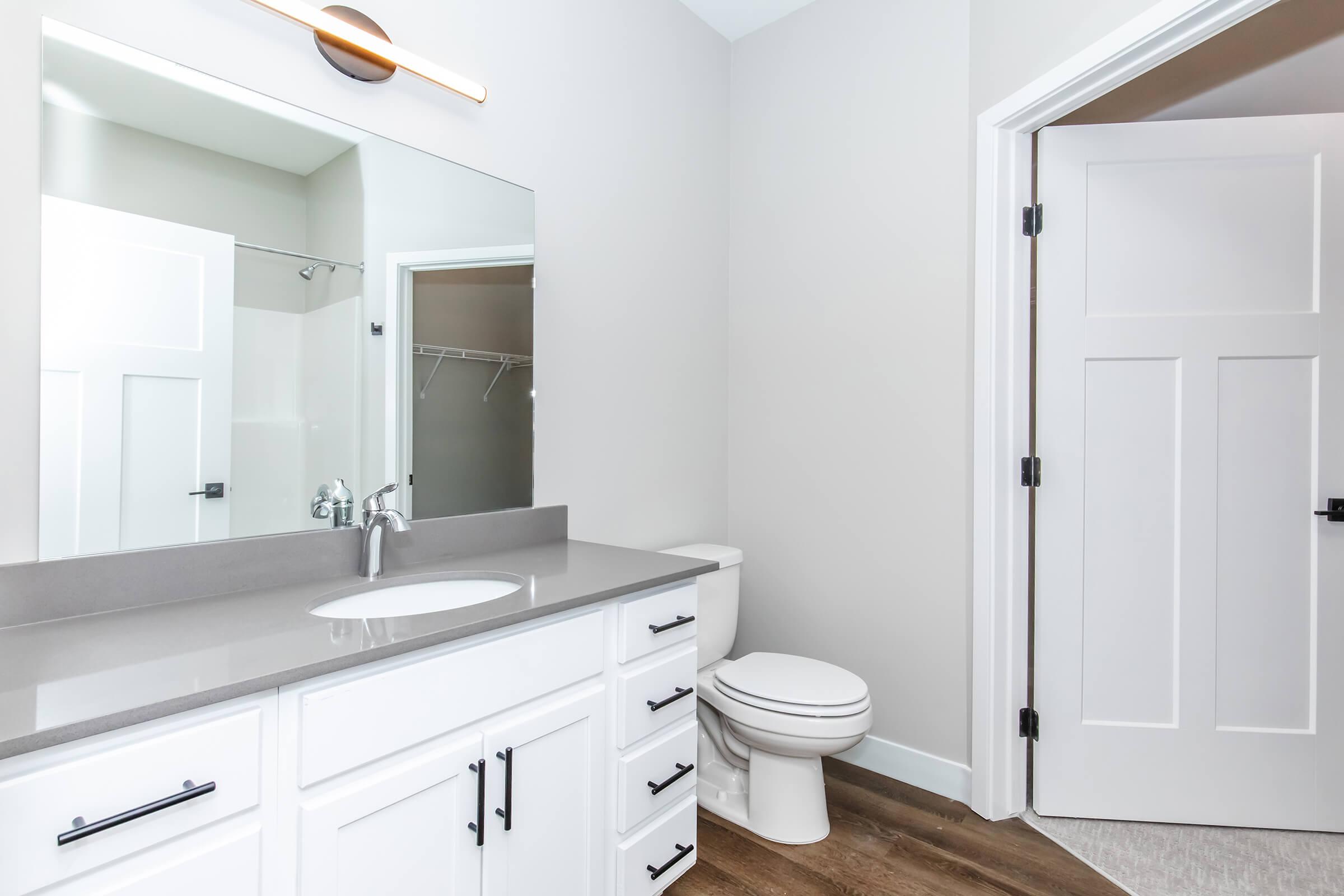 A modern bathroom featuring a large mirror above a gray countertop with a sink, white cabinetry, and stylish black hardware. There is a toilet on the side, and a doorway leading out of the room. The walls are painted in a light color, and the flooring is wood-like.