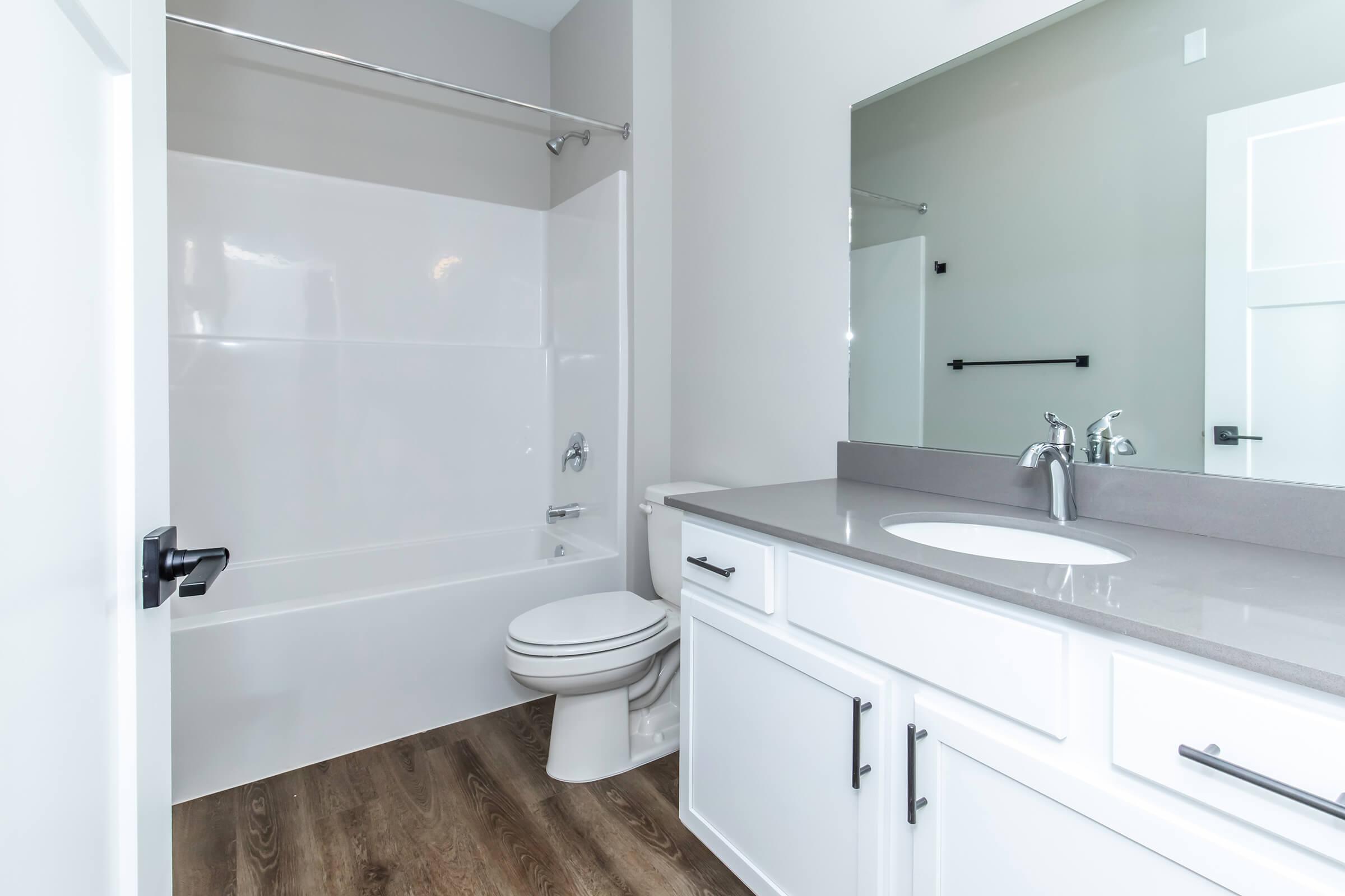 A modern bathroom featuring a white bathtub with a shower curtain, a toilet, and a sleek vanity with a sink and mirror. The walls are painted in a light gray tone, and the floor has a wood laminate finish, adding warmth to the space.