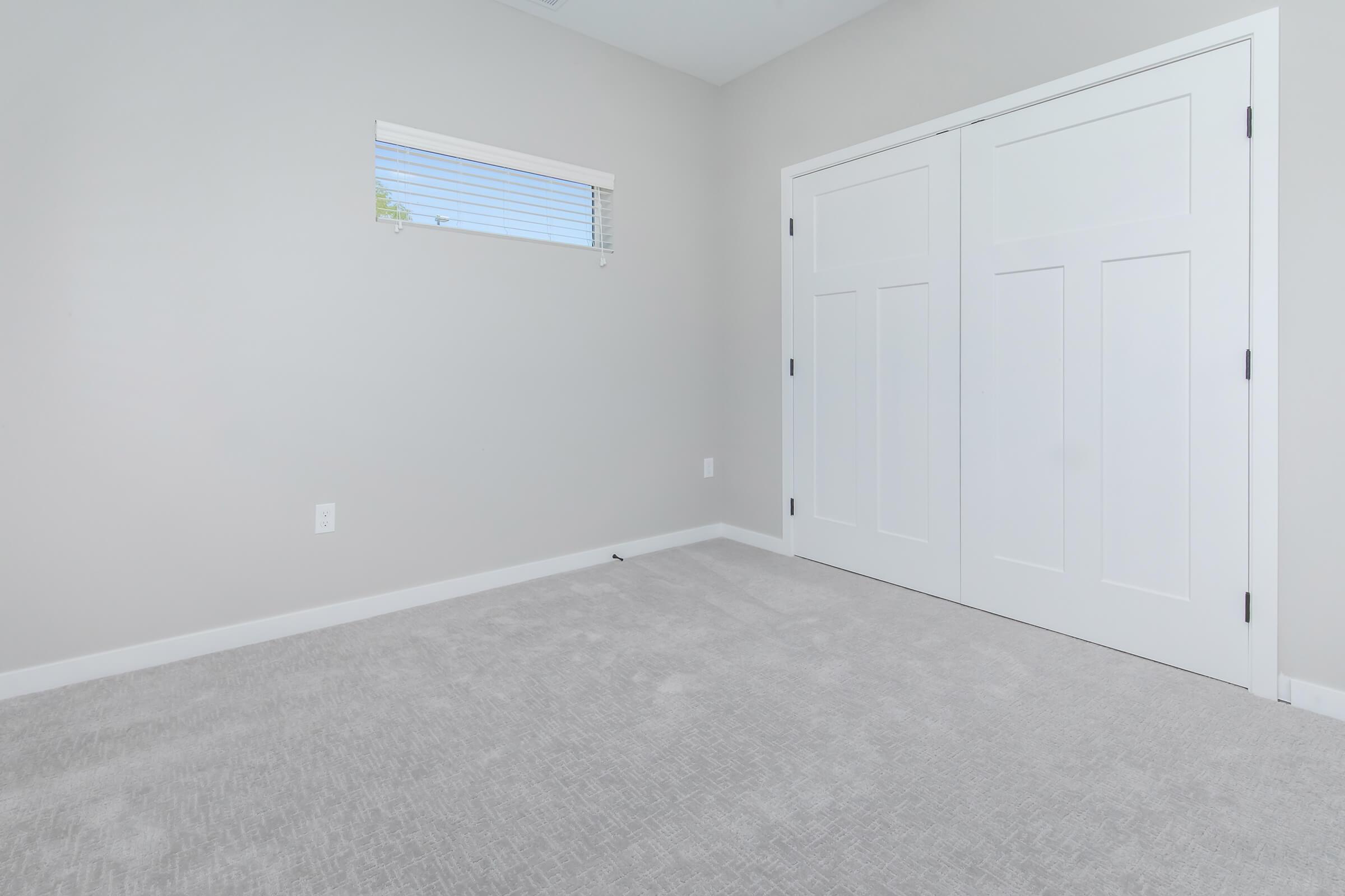 A light gray room with beige carpet, featuring a small window with horizontal blinds and two white-paneled doors. The walls are painted in a neutral shade, creating a bright and airy atmosphere.