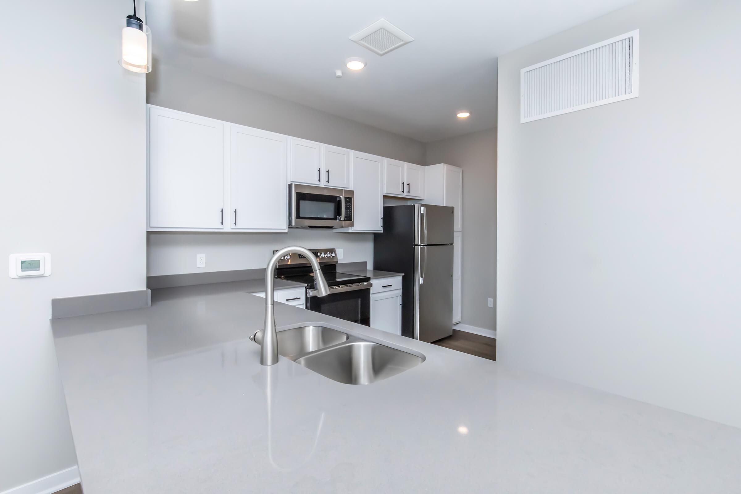 Modern kitchen with sleek white cabinets, stainless steel appliances, and a gray countertop. The design is open and minimalistic, featuring a double sink and pendant lighting. A refrigerator, microwave, and oven are visible, with a spacious, well-lit area ideal for cooking and entertaining.