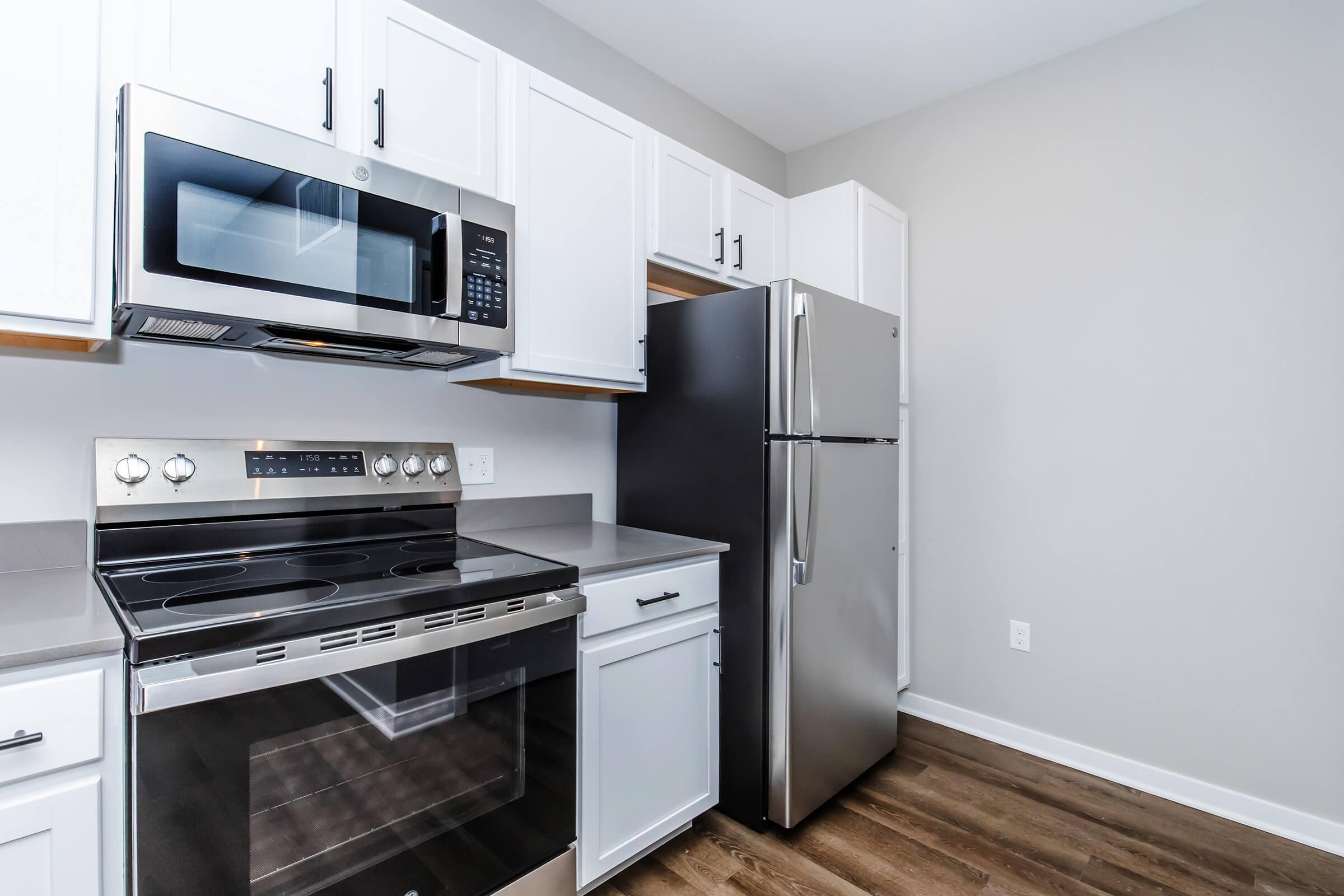 Modern kitchen featuring stainless steel appliances, including a microwave and oven, alongside a black refrigerator. The cabinets are white, and the countertops are gray. The flooring is a warm wood tone, creating a contemporary and inviting space.