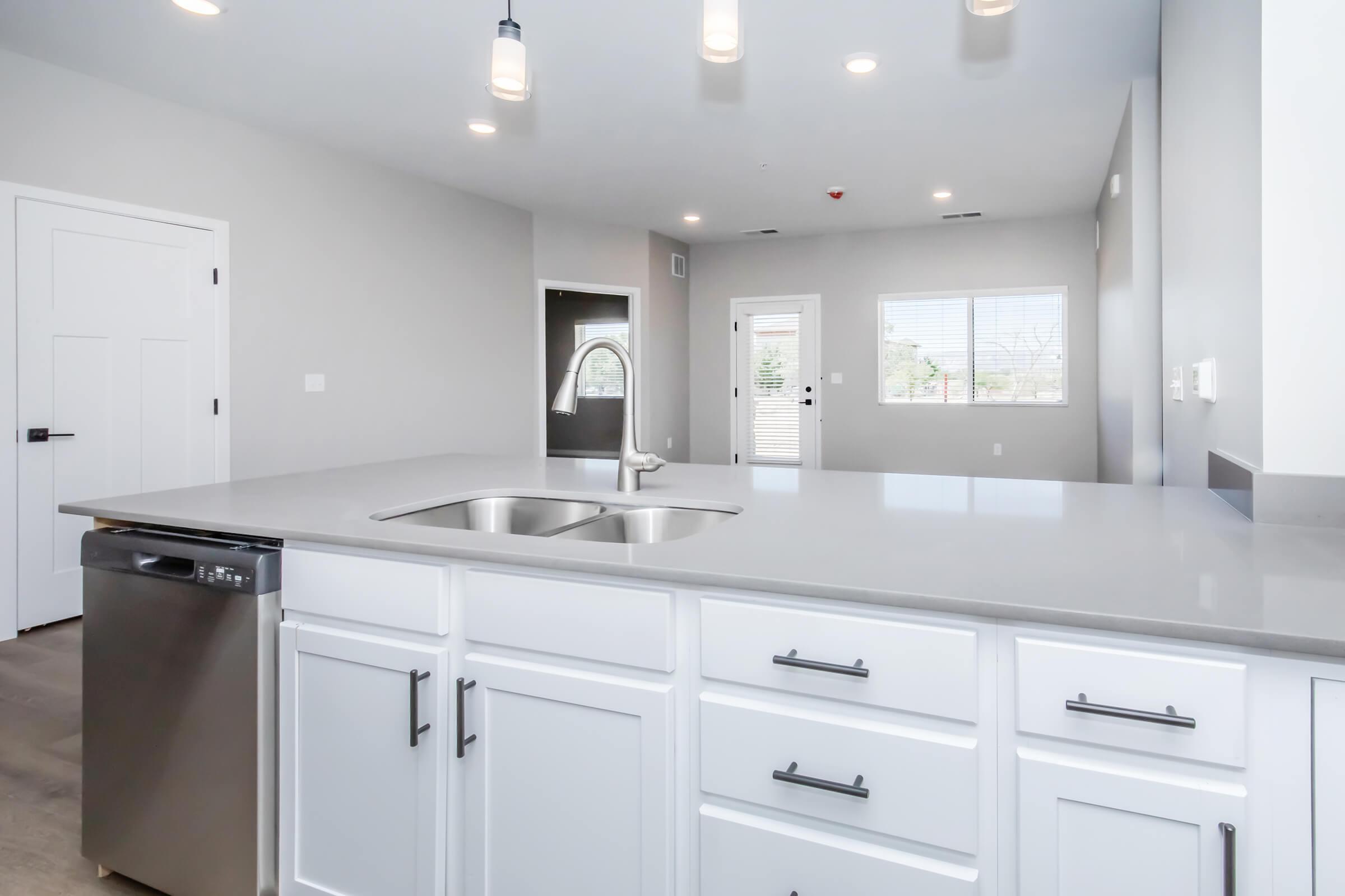 Modern kitchen interior featuring a large island with a double sink, stainless steel dishwasher, and sleek white cabinetry. The walls are painted light gray, and pendant lights hang from the ceiling. A view of an open living area with natural light and windows can be seen in the background.