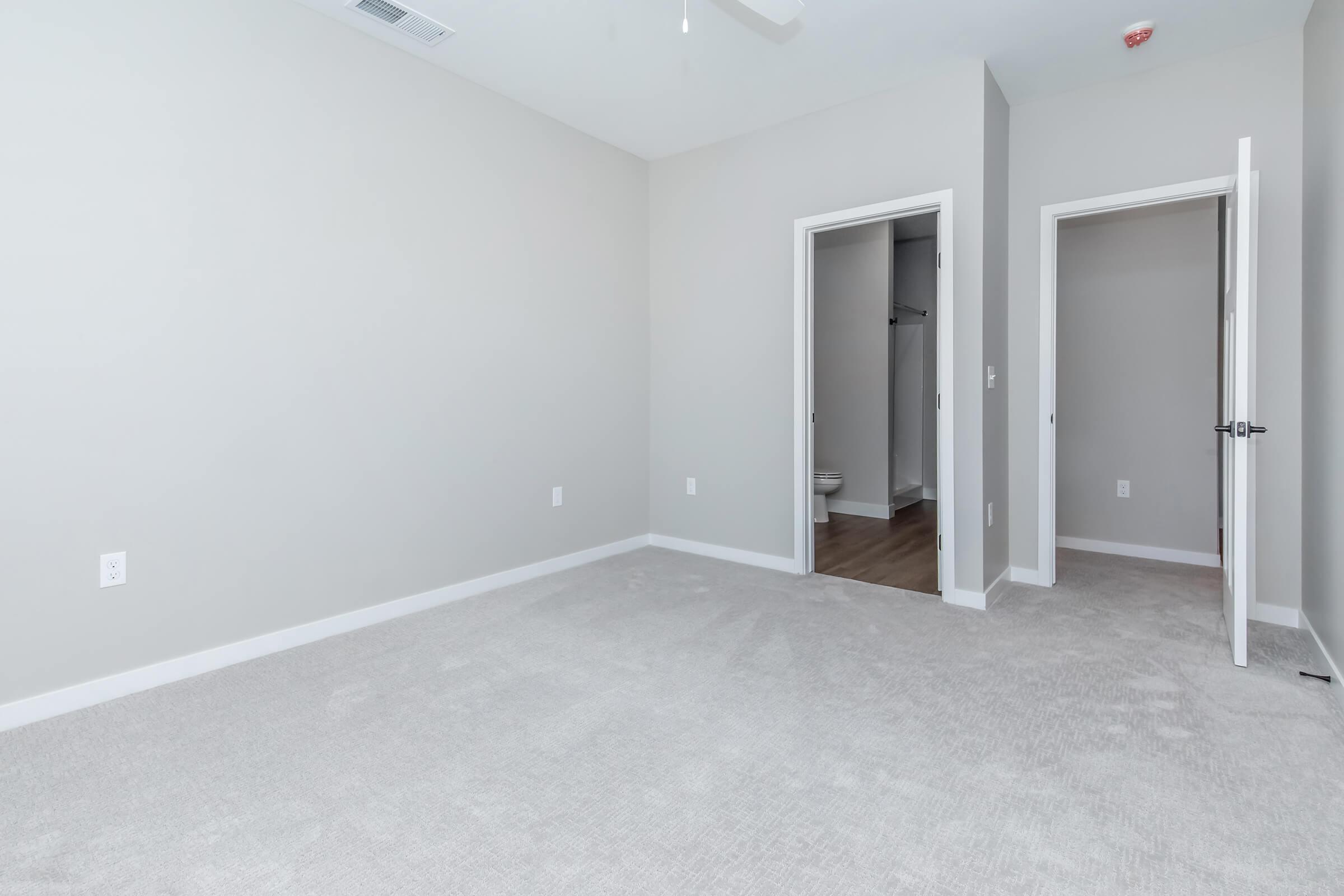 A spacious, empty room with light gray walls and carpet. There are two doorways leading to adjacent rooms, one of which has a visible toilet area. The overall ambiance is clean and modern, with a ceiling fan fixture and ample natural light.