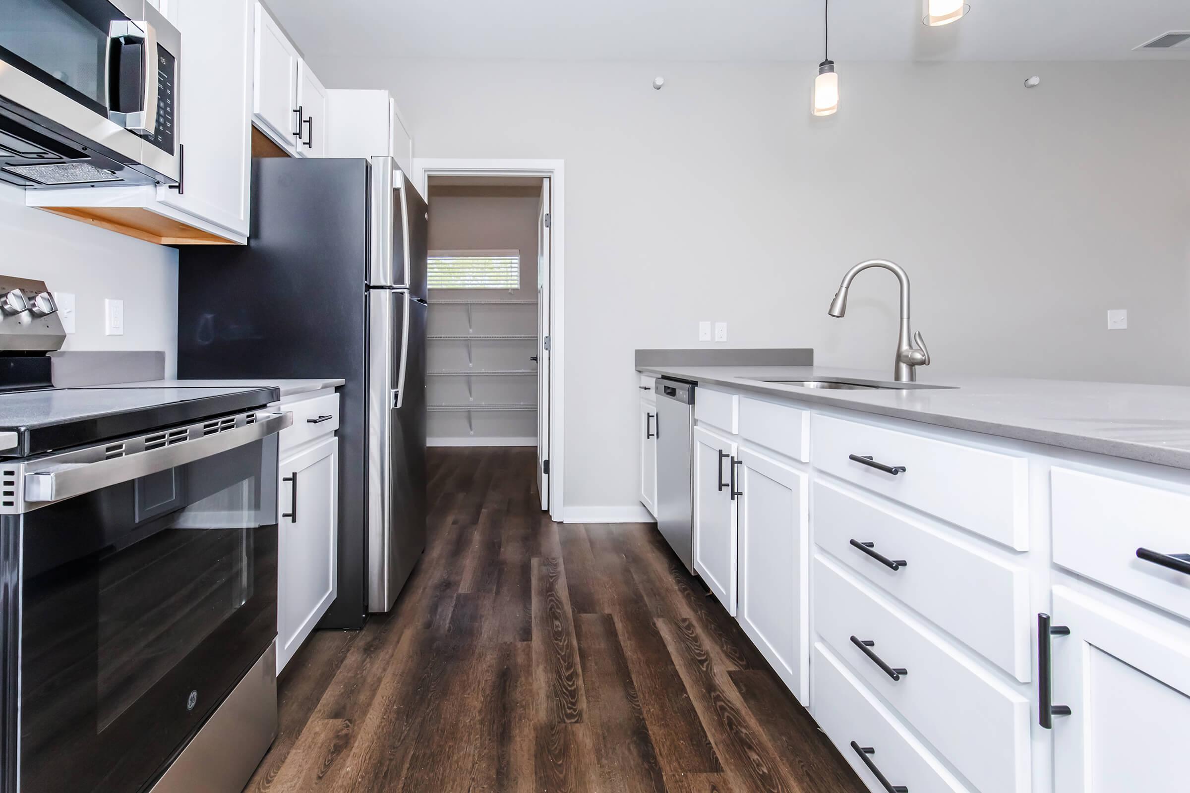 A modern kitchen featuring stainless steel appliances, including a microwave and refrigerator. The kitchen has white cabinetry with black handles, a large sink with a sleek faucet, and a spacious island. The flooring is a dark wood, and there is a pantry visible through an open door. The walls are painted light gray.