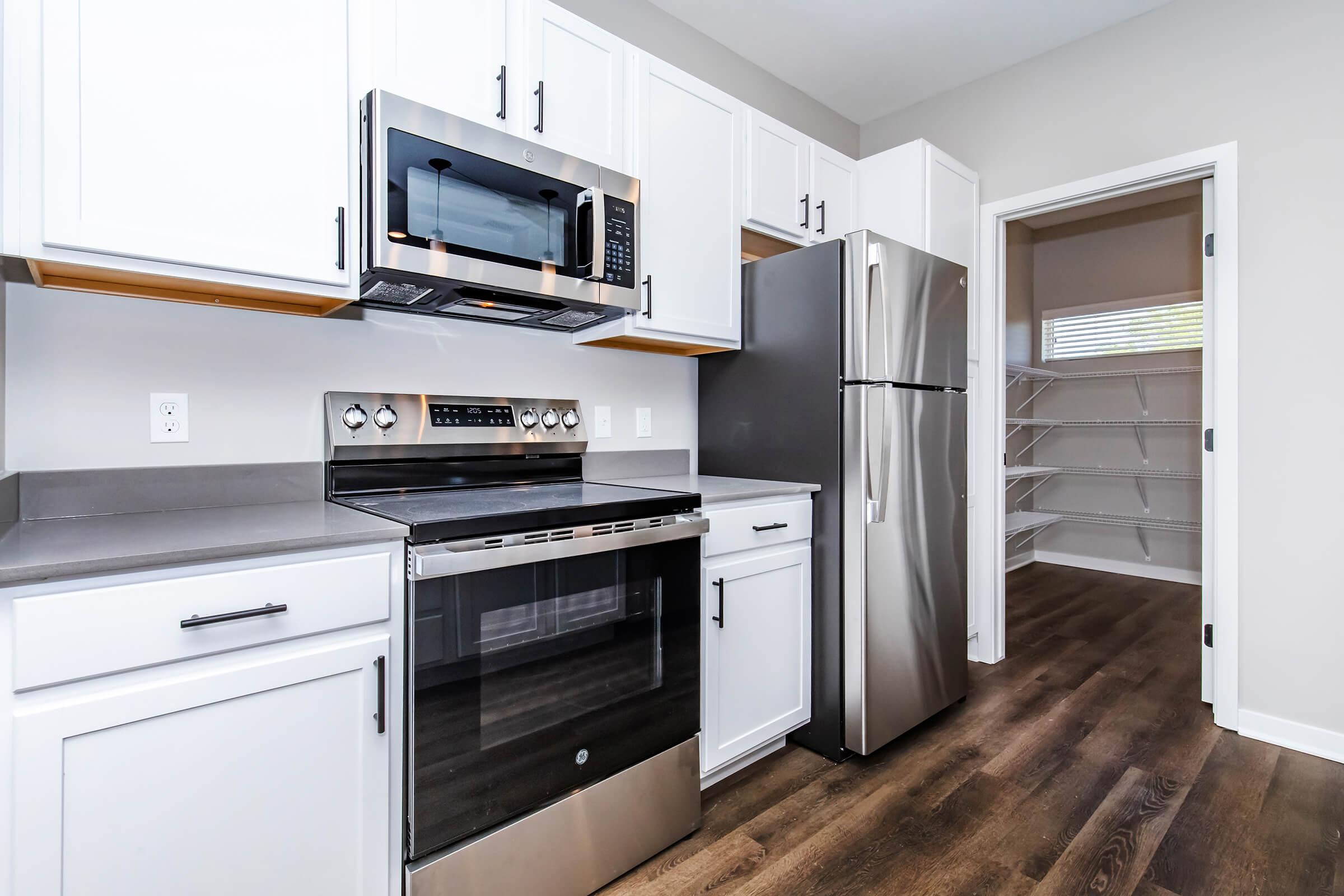 Modern kitchen featuring stainless steel appliances, including a microwave, oven, and refrigerator. The cabinets are white with a sleek design, complemented by gray countertops. A pantry is visible in the background, with open shelving for storage, and the floor is finished in wood-like laminate.