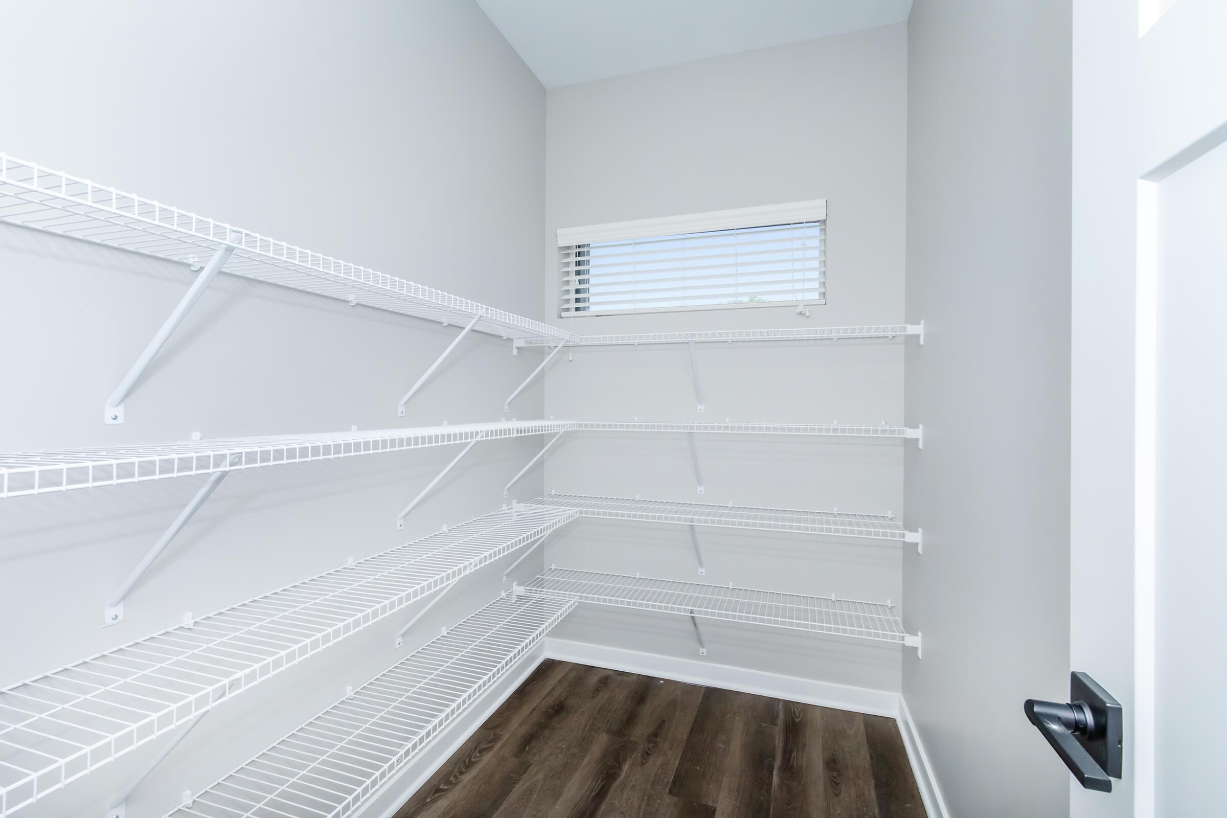 A spacious, empty pantry with white wire shelves mounted on light gray walls. Natural light streams in through a small window with blinds, and the floor features dark wood laminate. The room is uncluttered and ready for storage.