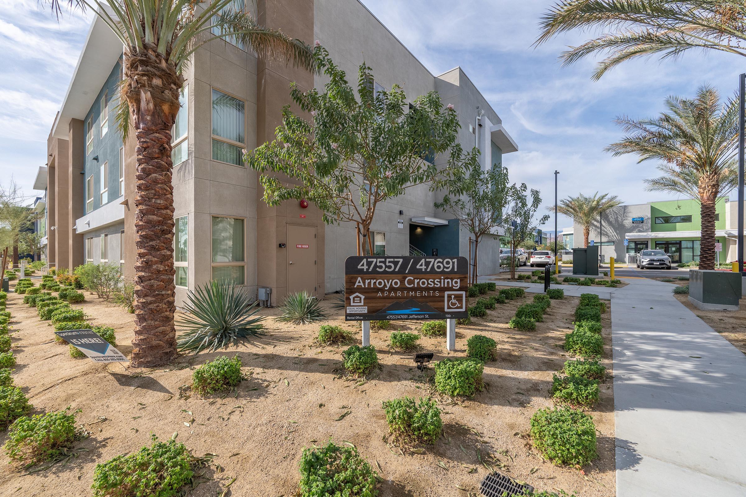 A modern apartment building with the sign "Arroyo Crossing" displayed prominently in front. The area features landscaped shrubs, palm trees, and well-maintained pathways. Sunny weather with a clear sky is visible, and nearby structures can be seen in the background.
