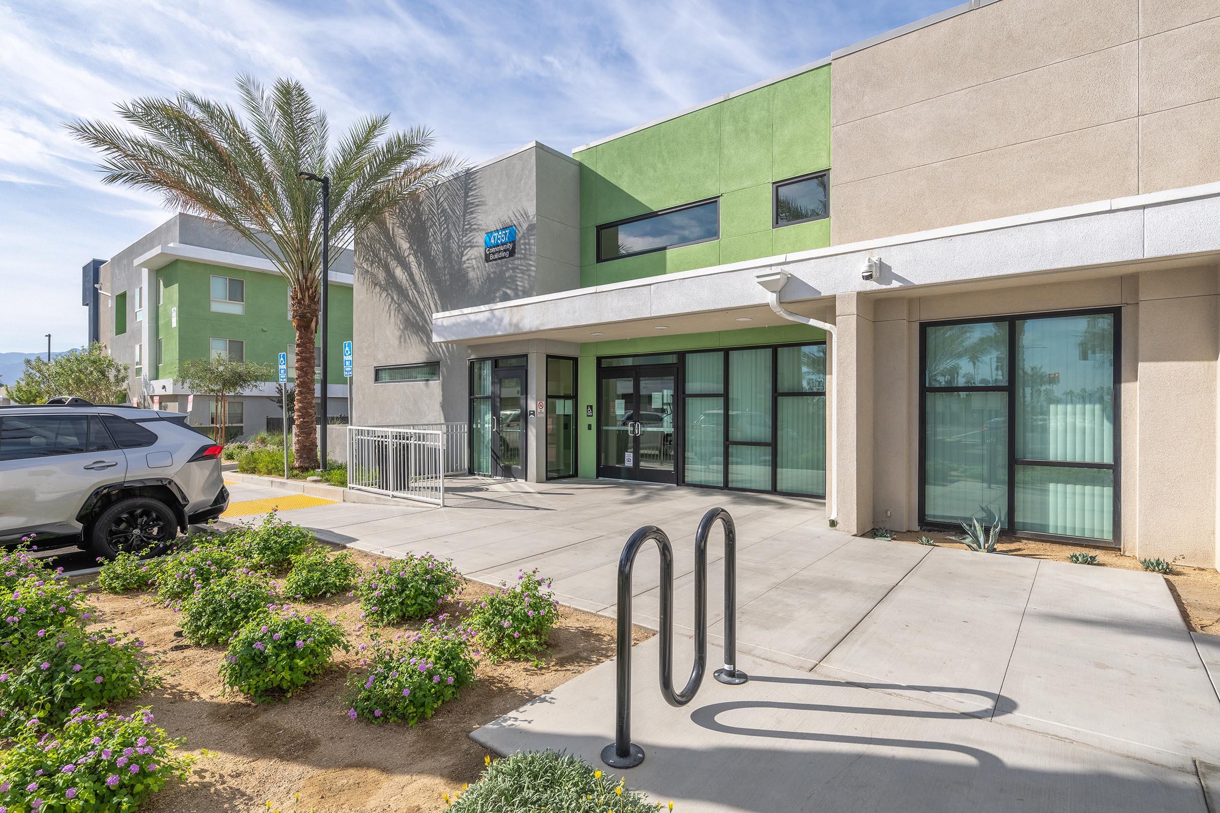Modern building entrance with large glass doors flanked by palm trees and low shrubs. A light-colored vehicle is parked nearby. The facade features a mix of green and beige colors, with a sidewalk area leading to the entrance. Bike racks are visible in the foreground.