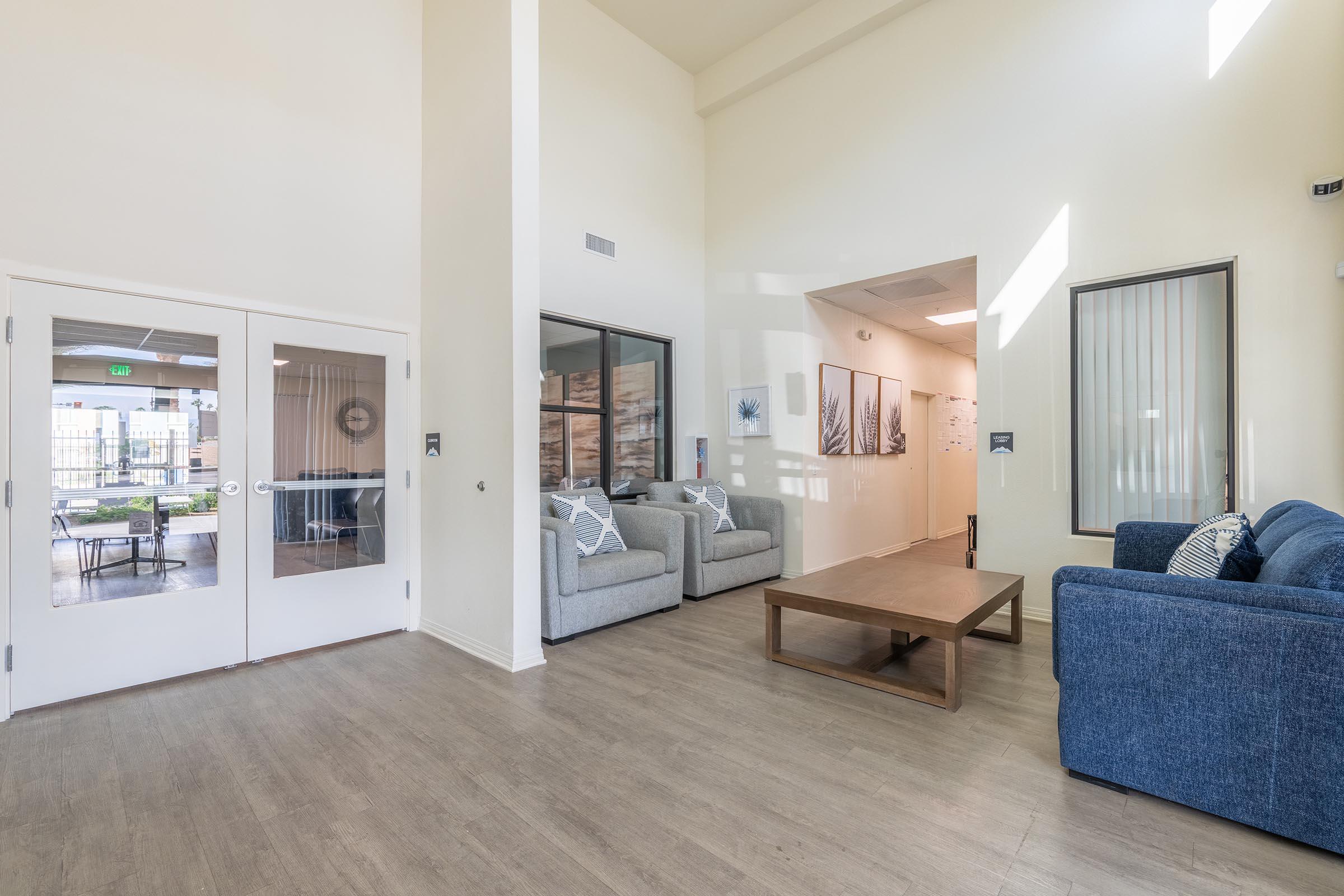 A bright, modern living space featuring two gray sofas and one blue sofa arranged around a wooden coffee table. The walls are painted light colors, and large windows allow natural light to fill the room. In the background, an entrance leading to another area is visible.