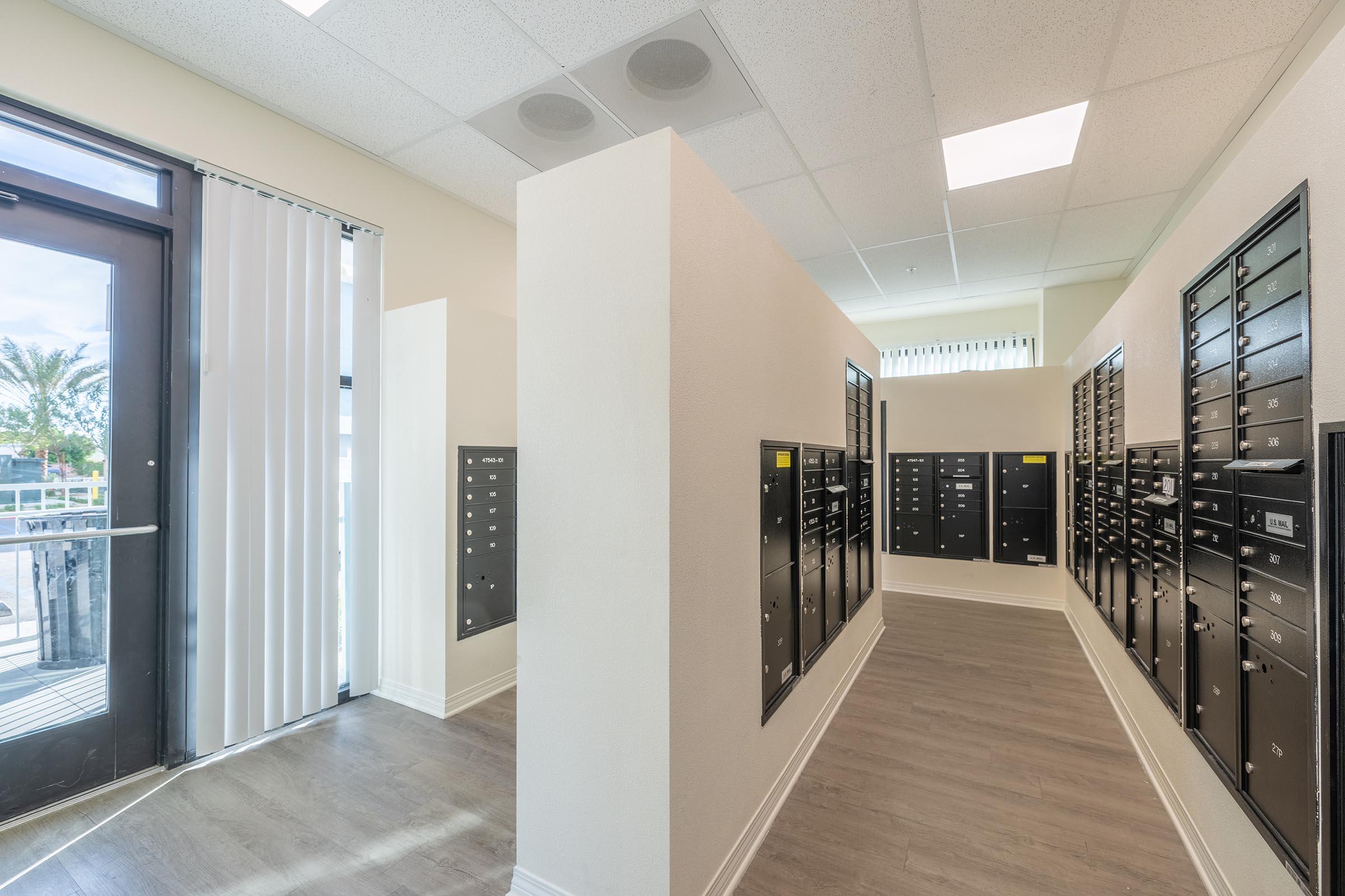 Interior view of a mailroom with two rows of black mailboxes lining the walls. The space is well-lit with overhead lighting, featuring a doorway to the outside. The floors are light wood, creating a modern and organized appearance. There are palm trees visible outside through the doorway.