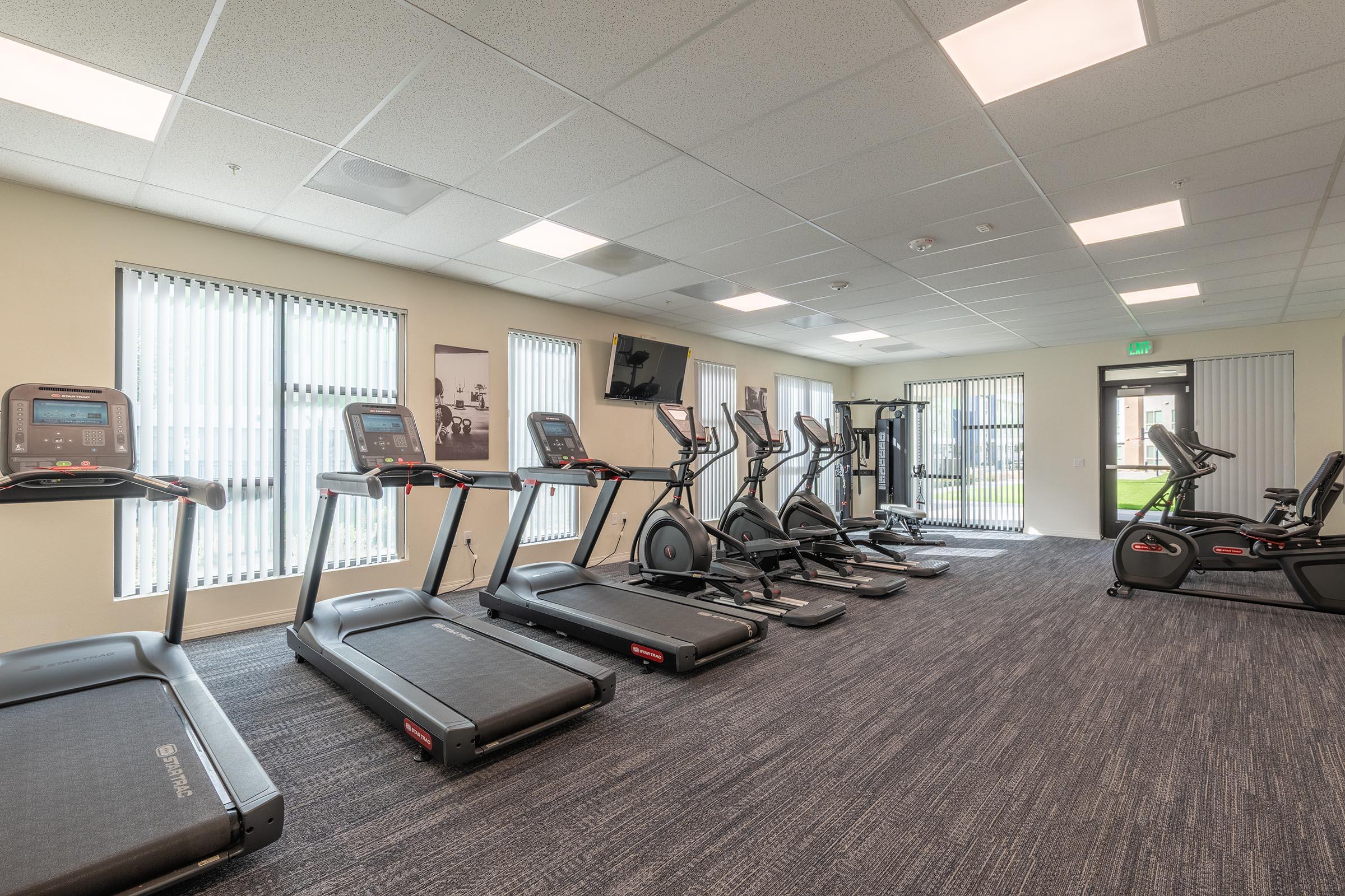 A modern gym interior featuring several treadmills and elliptical machines arranged neatly. Large windows allow natural light to fill the room, and there is a wall-mounted TV for entertainment. The facility has a clean, inviting appearance with carpeted flooring and an open layout.