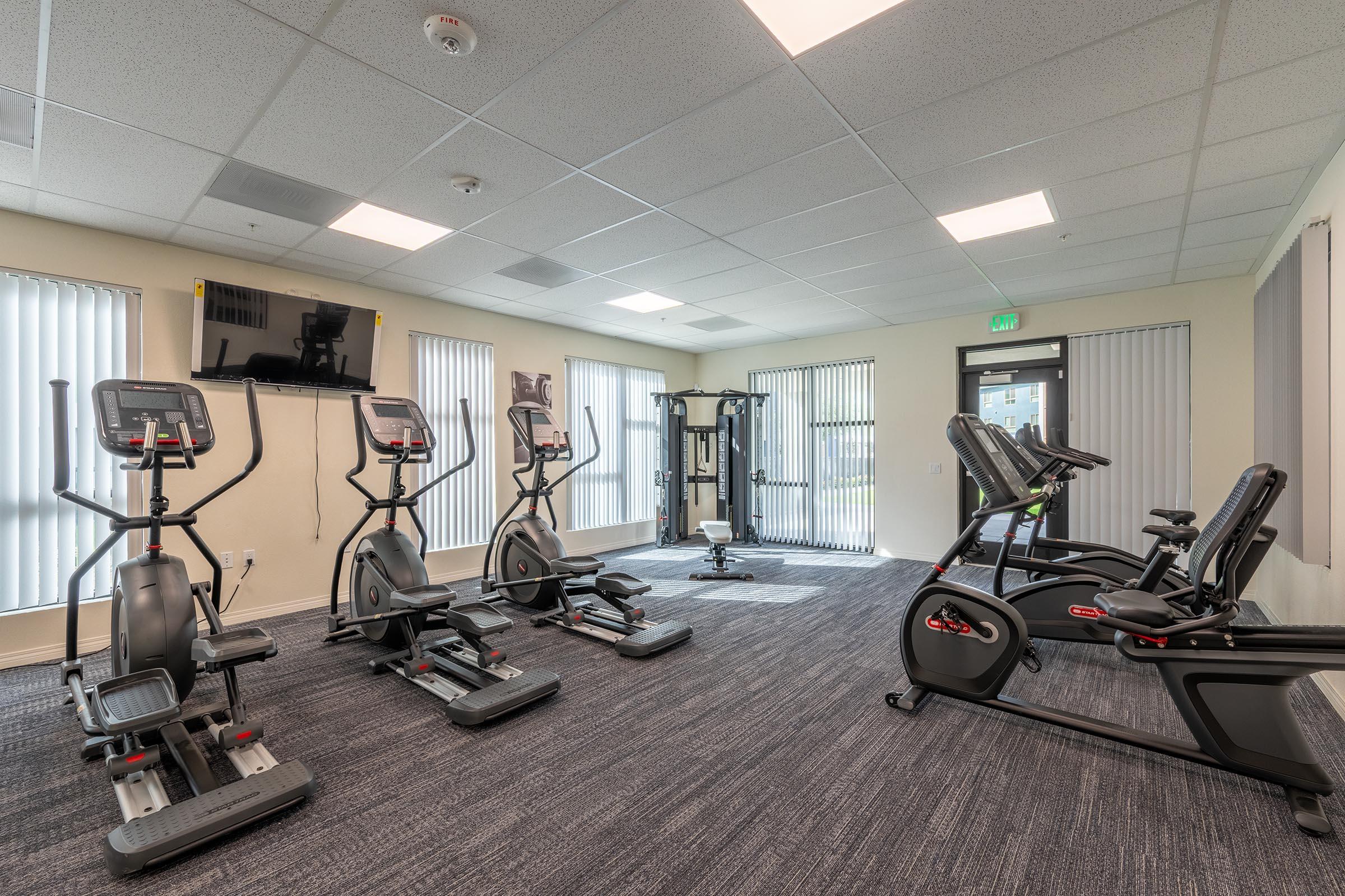 A well-lit gym interior featuring several exercise machines, including elliptical trainers and a recumbent bike. Large windows with blinds allow natural light, and there are wall-mounted televisions. The floor is covered with dark carpet, creating a professional workout environment.