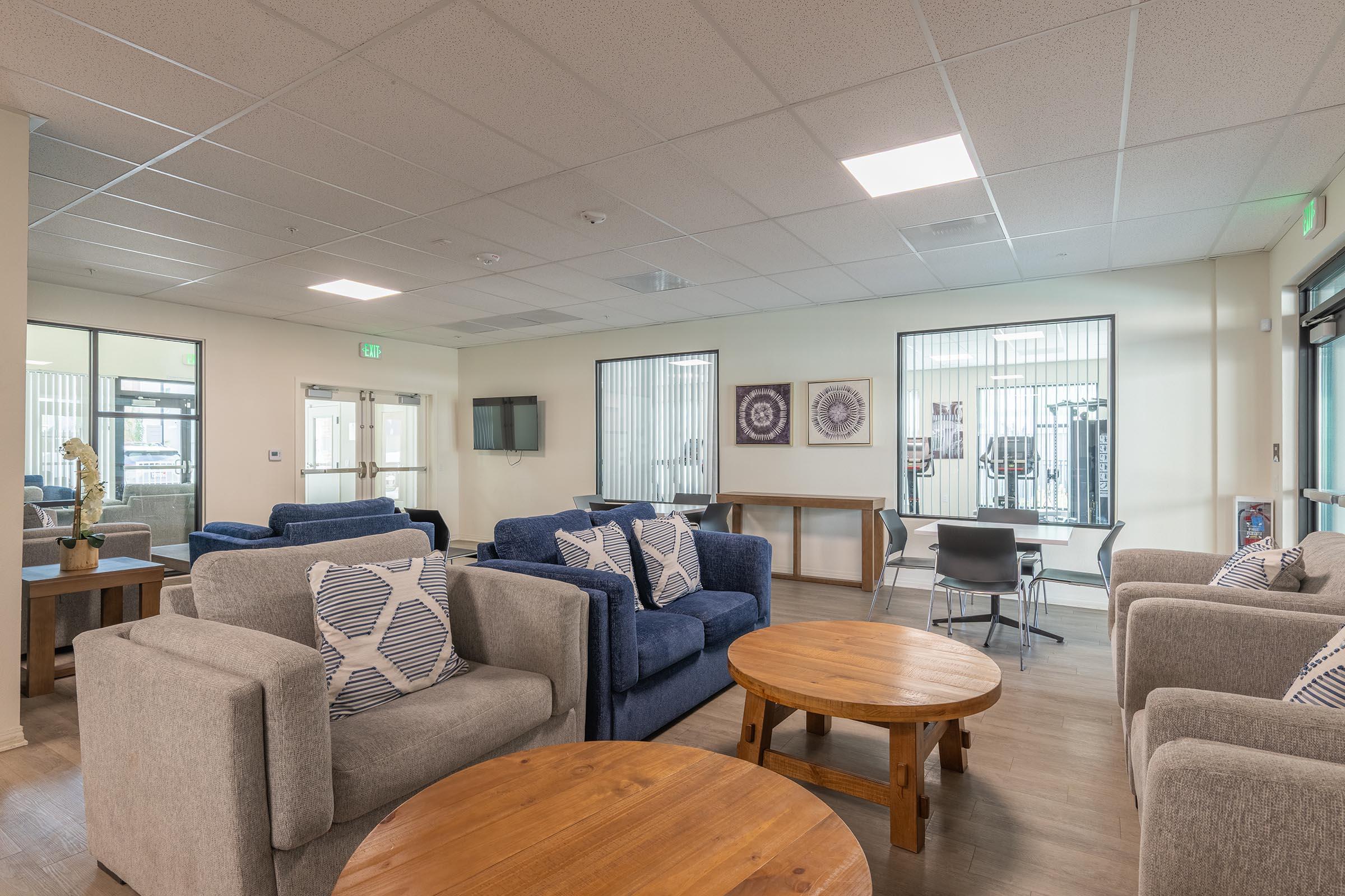 A modern lounge area featuring several comfortable sofas with decorative pillows, wooden coffee tables, and a simple chair. The space is well-lit with large windows, and there's a TV mounted on the wall. Fitness equipment is visible through a glass partition in the background.