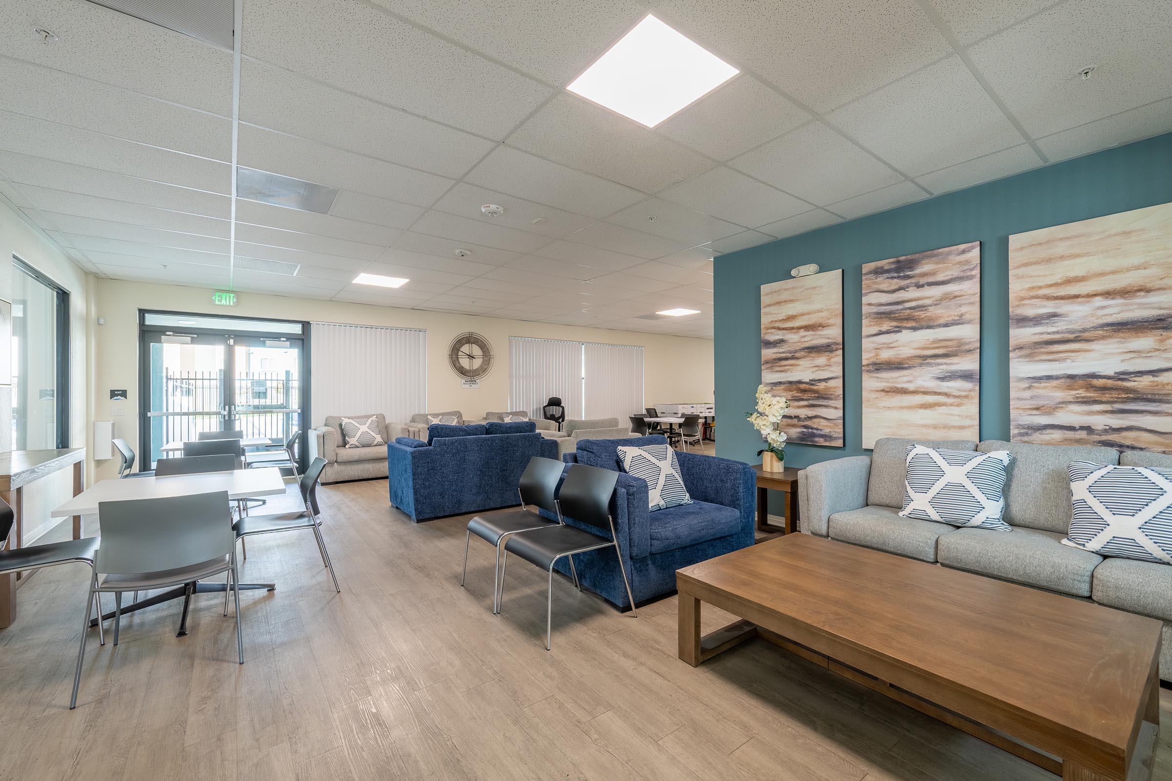 A spacious lounge area featuring two blue sofas and a light gray couch, arranged around a wooden coffee table. Wall art with abstract designs hangs on a blue accent wall. There are several tables and chairs for seating, with large windows providing natural light.