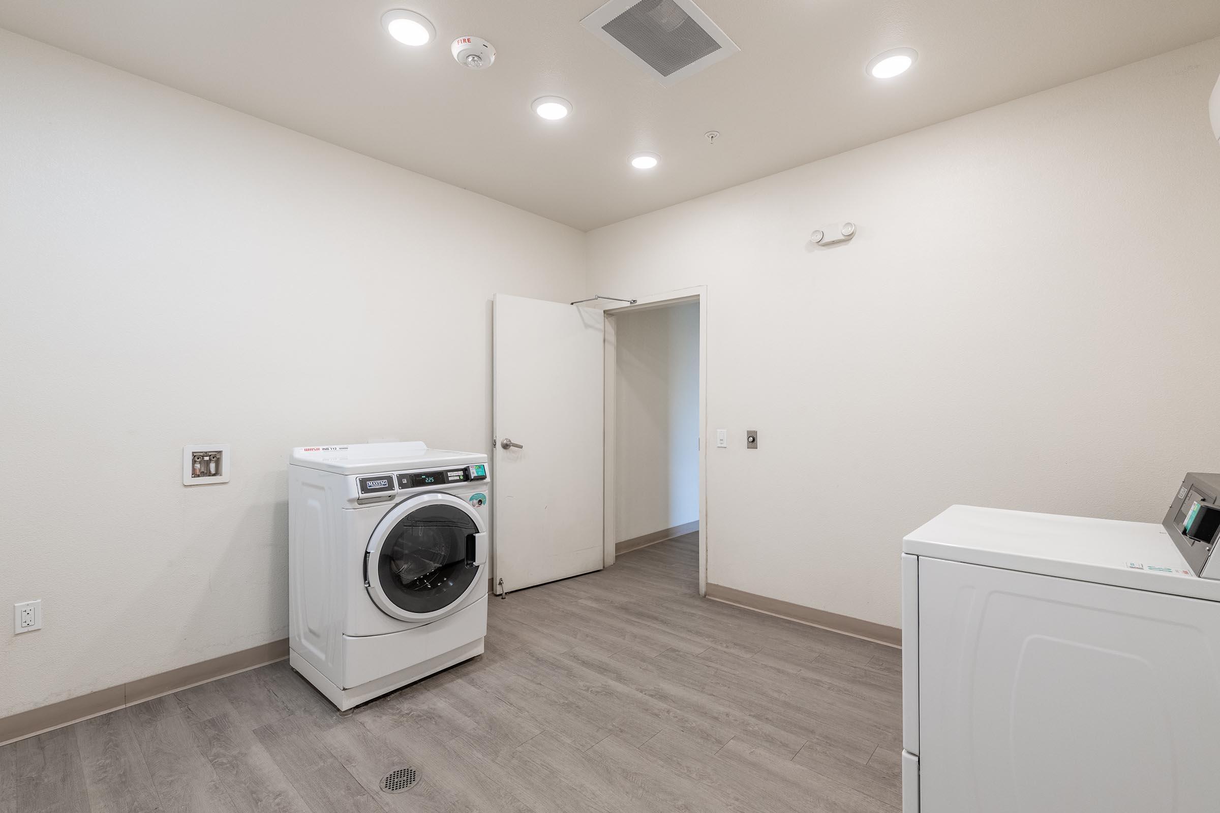 A clean, modern laundry room featuring a front-loading washing machine on the left and a dryer on the right. The walls are painted light-colored, and there is a door leading to another room. The floor is a light wood laminate, and ceiling lights provide bright illumination.