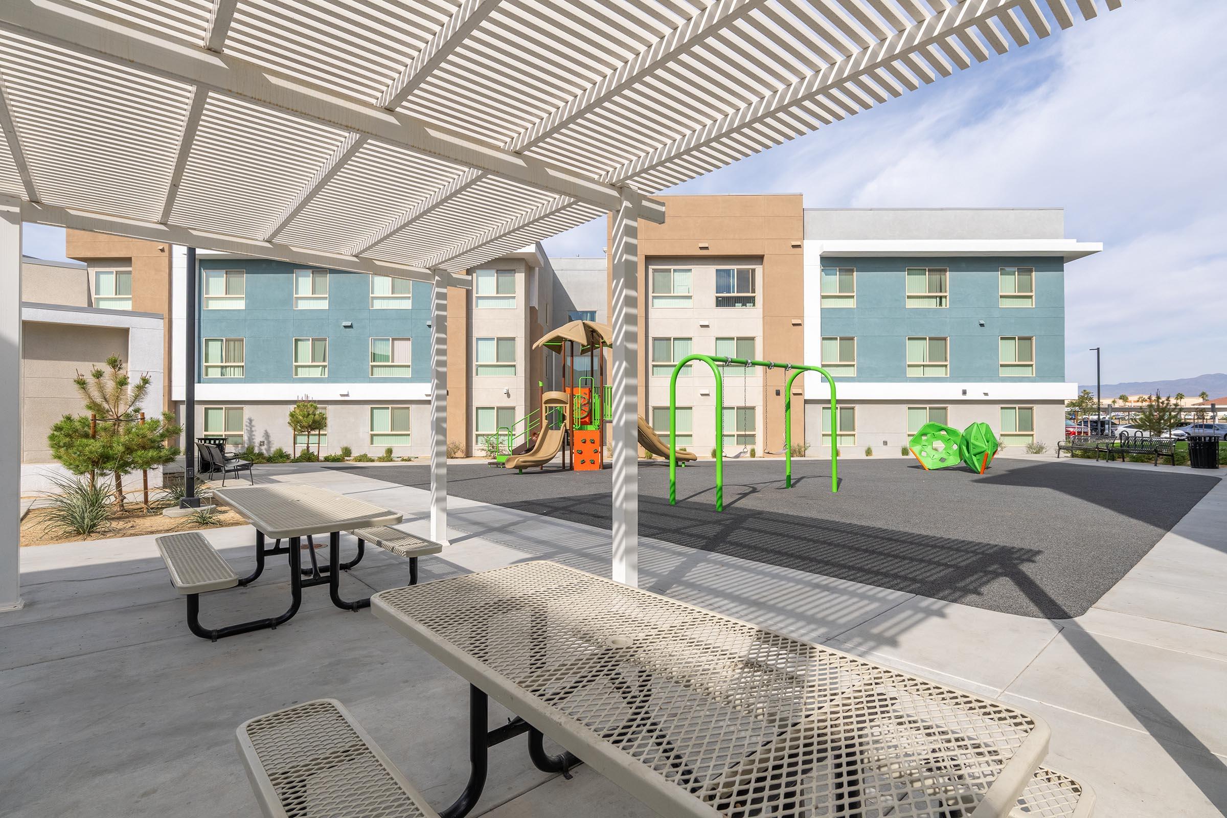 A playground area features colorful equipment including slides and climbing structures, surrounded by grassy patches. In the foreground, there is a shaded picnic area with metal tables and benches, set against the backdrop of modern residential buildings. The sky is partially cloudy, indicating a bright day.