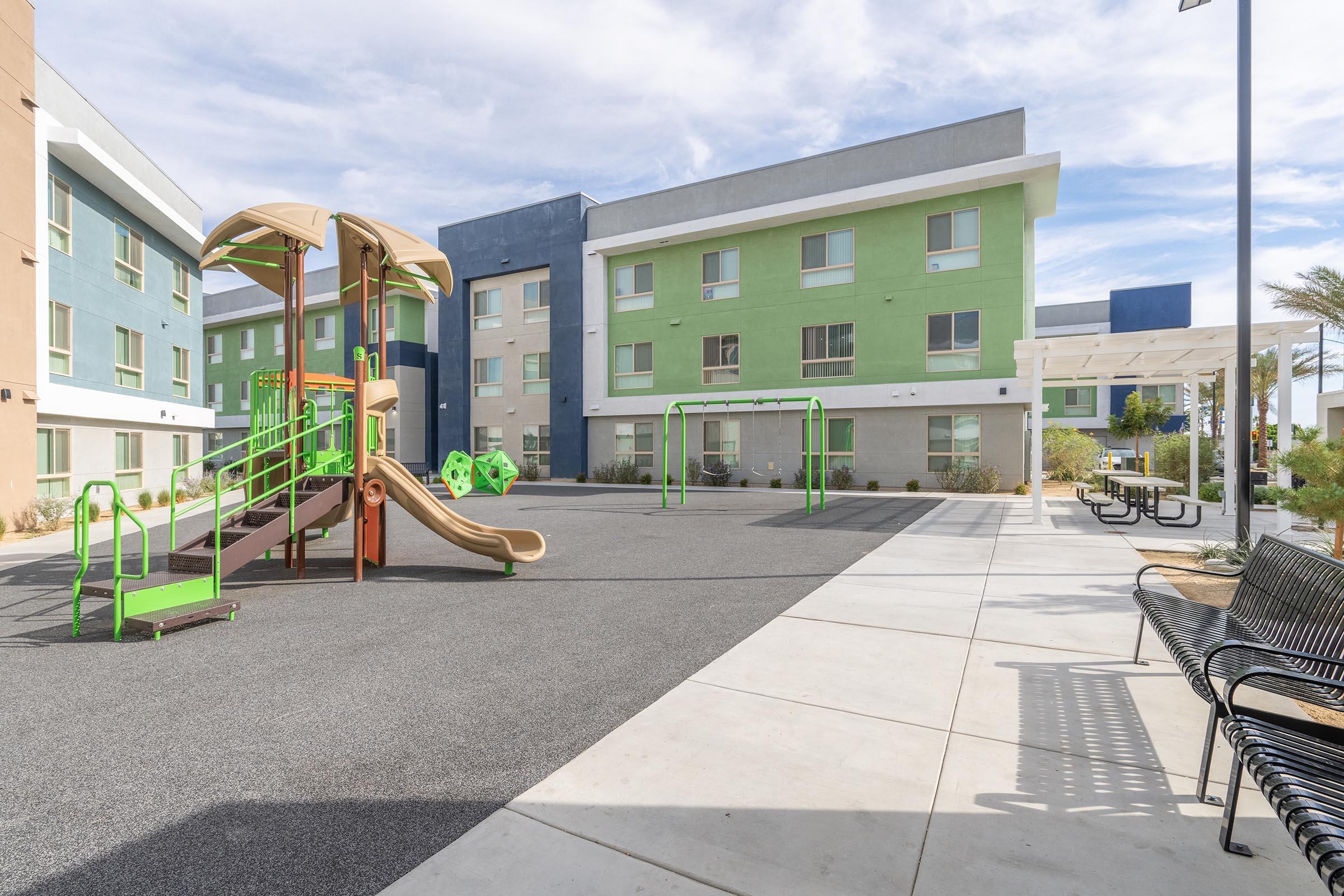 A playground featuring a wooden structure with slides, climbing areas, and swings is situated in a spacious outdoor area. Surrounding the playground are modern buildings painted in green and blue, with shaded seating available nearby and a clear blue sky overhead.