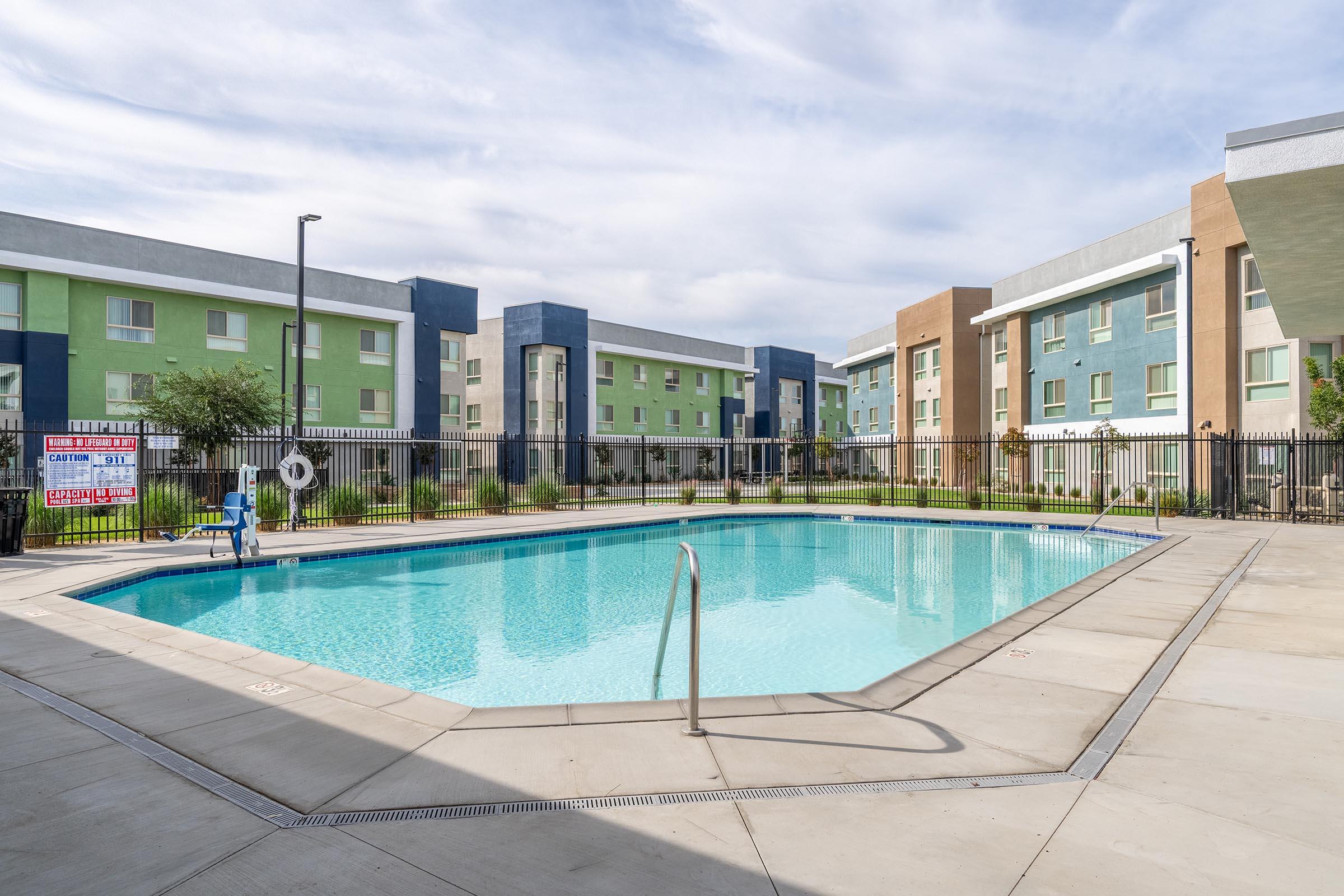 A clear swimming pool surrounded by a concrete deck, within a fenced area. Apartment buildings in shades of green and brown are visible in the background under a partly cloudy sky. A pool ladder is positioned at the edge, and there are lounge chairs nearby.