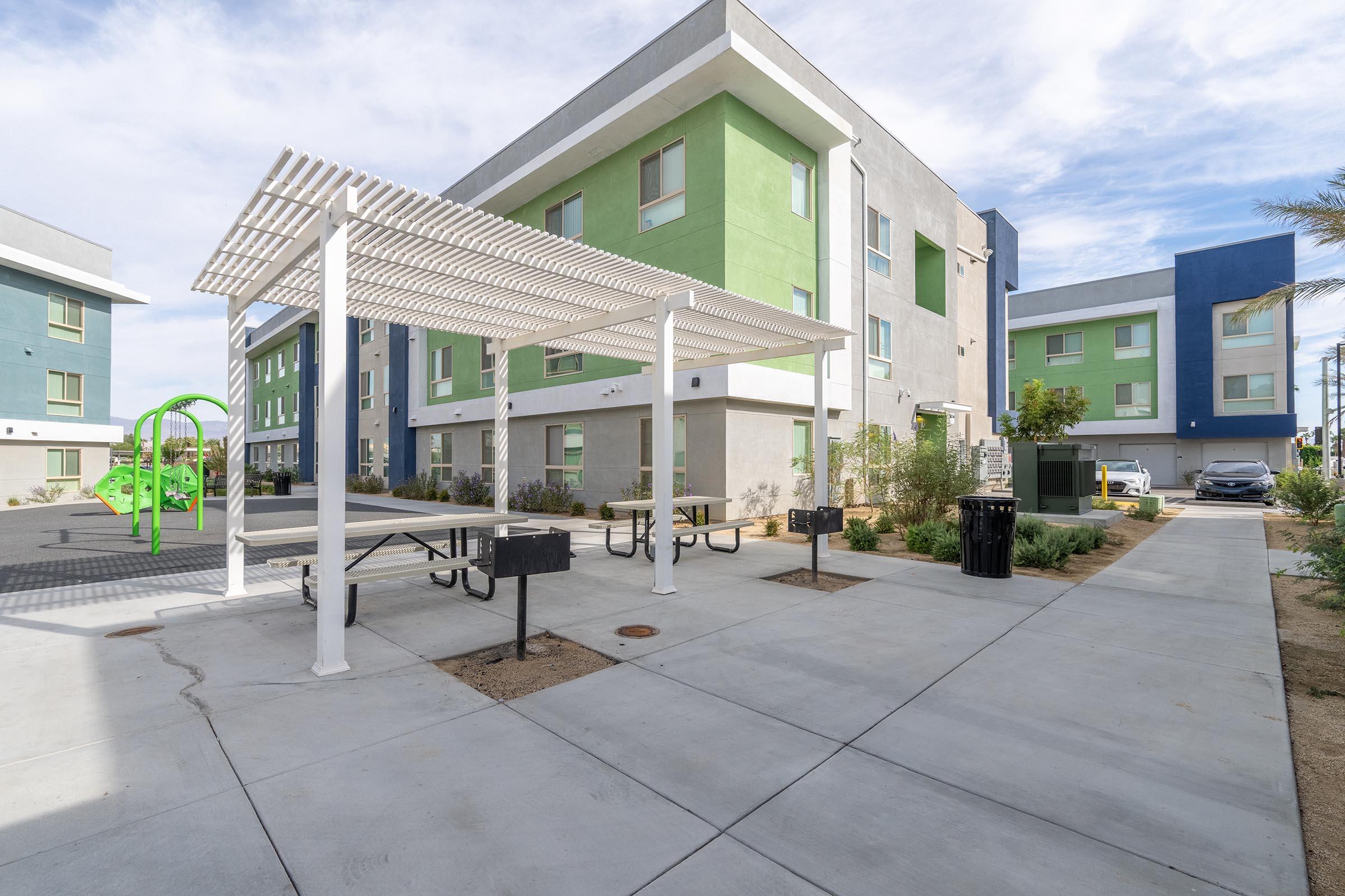Modern residential complex with green and blue buildings. In the foreground, a shaded picnic area with tables and grills is visible, alongside a playground with green equipment. The pathway leads to a clean, landscaped area, with additional parking in the background. Clear skies enhance the setting.