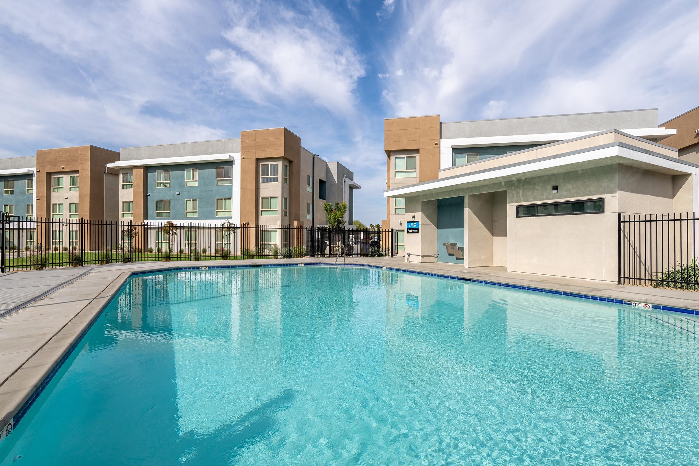 A clear swimming pool surrounded by a concrete deck, with lounge chairs nearby. In the background, there are two modern apartment buildings featuring a mix of blue and beige facades under a partly cloudy sky. A fenced area encloses the pool for privacy and safety.