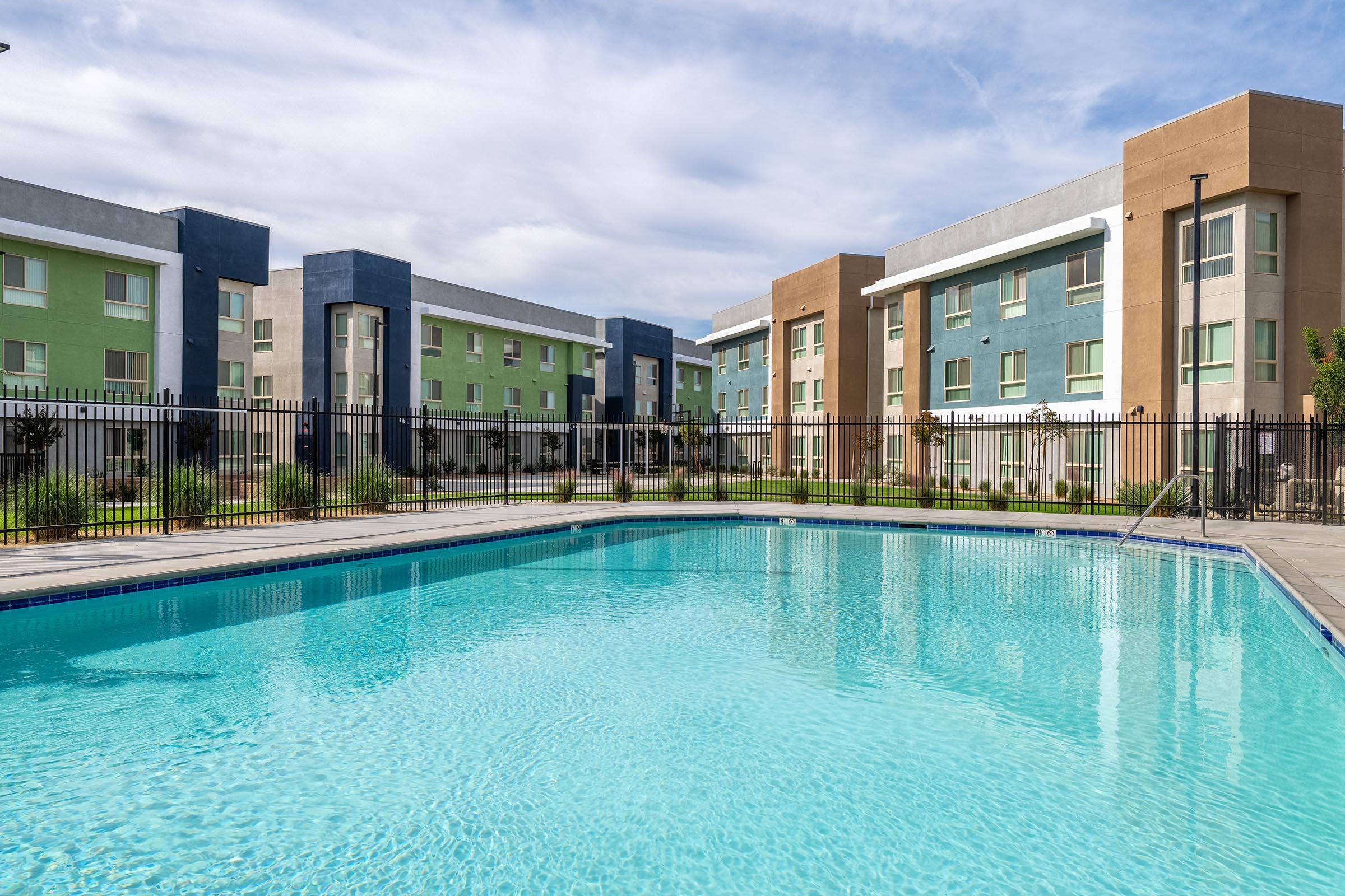 A clear blue swimming pool in the foreground, surrounded by a fenced area. In the background, modern apartment buildings featuring a mix of pastel colors, such as green, blue, and tan. The sky is partially cloudy, creating a bright and inviting atmosphere.