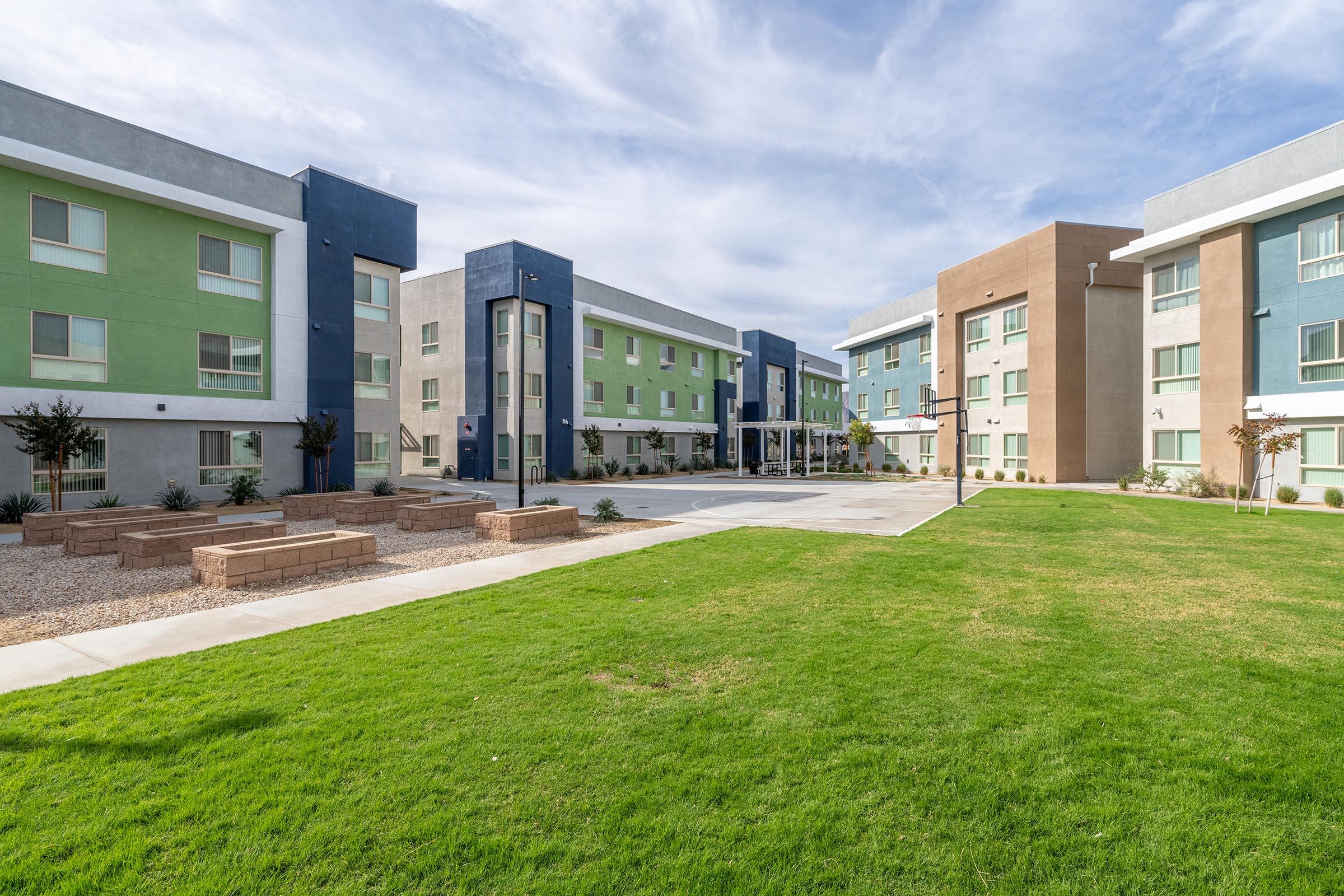 Modern apartment complex with two buildings featuring colorful facades in green, blue, and brown. A landscaped courtyard with neatly trimmed grass and stone seating areas is visible in front of the buildings. The sky is partly cloudy, adding a bright ambiance to the scene.