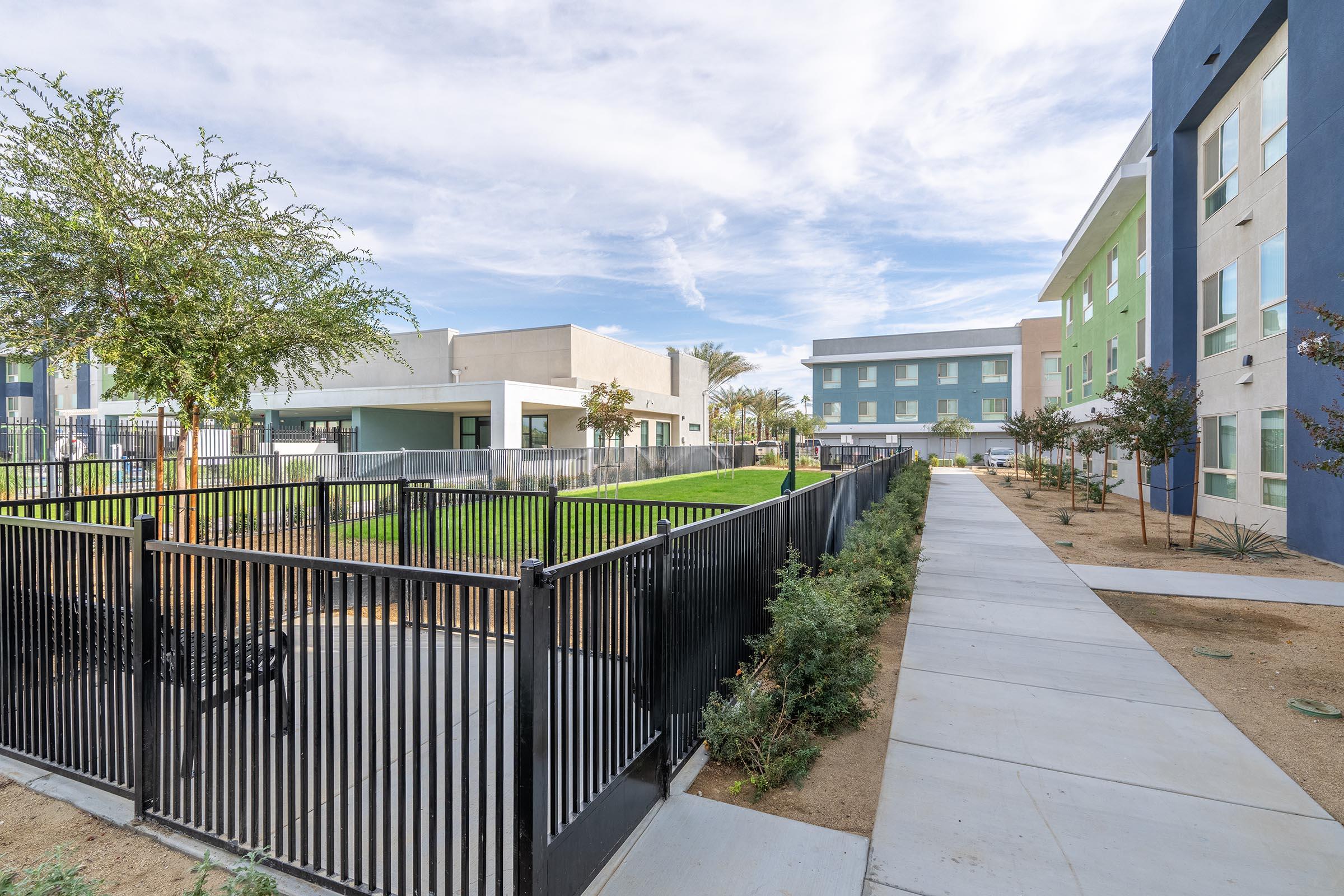 A landscaped area featuring a grassy lawn enclosed by a black fence, with a concrete pathway running alongside. Modern buildings in various colors are visible in the background, under a partly cloudy sky. Trees and shrubs are planted along the pathway, contributing to the outdoor aesthetic.