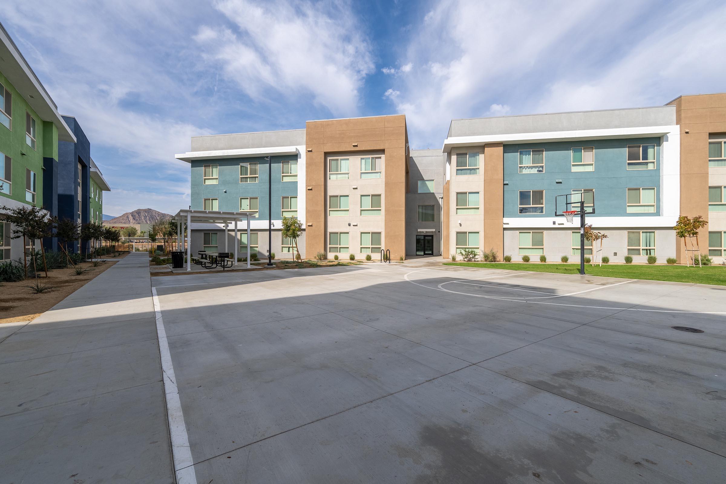 Modern two-story residential buildings in green and beige, set in a spacious outdoor area with a basketball hoop and paved court. The sky is bright blue with scattered clouds, creating a welcoming and bright environment.