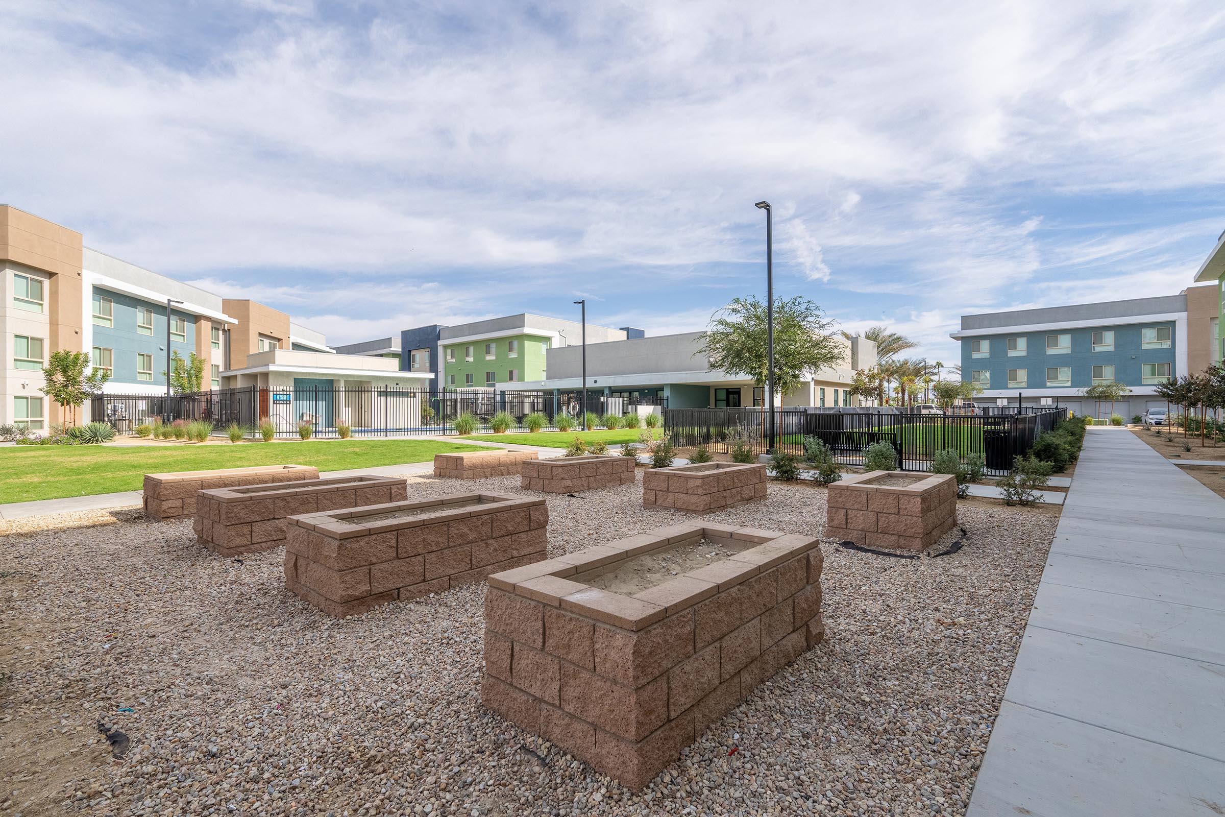 A landscaped area featuring several rectangular stone planters surrounded by gravel. In the background, there are modern multi-story buildings with a mix of light-colored façades and green accents. The scene is under a partly cloudy sky, creating a tranquil outdoor setting.