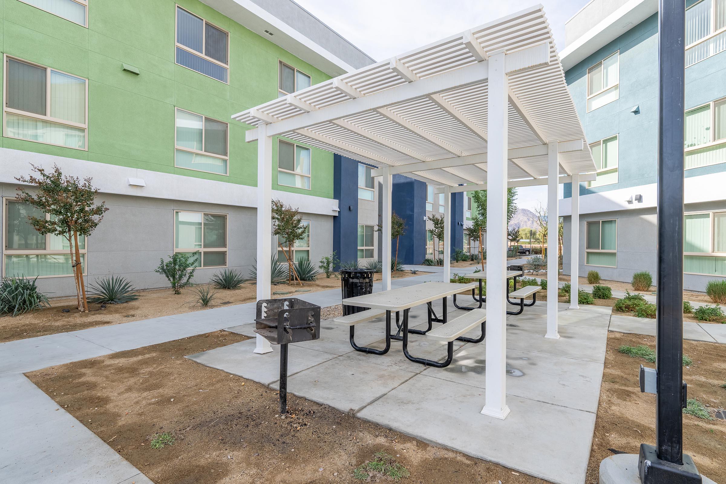 A modern outdoor seating area featuring a white pergola with picnic tables, surrounded by low shrubs and plants. Nearby, a barbecue grill is available for use. The background includes colorful building exteriors in green and blue. The area is designed for relaxation and social gatherings.