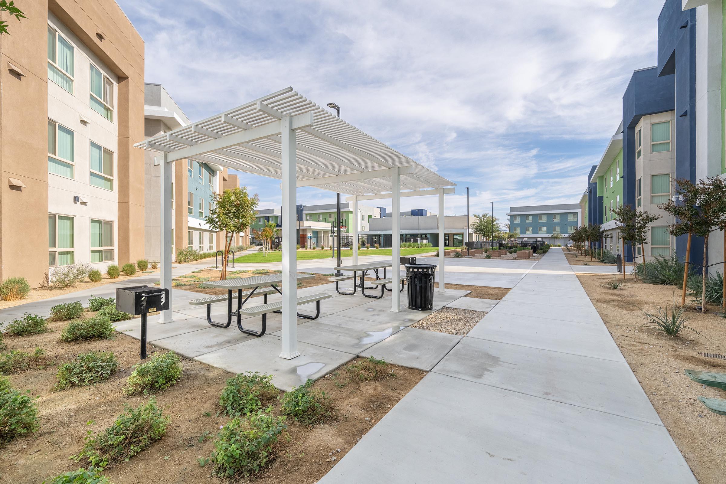 A modern outdoor area featuring a picnic table under a white pergola, surrounded by green shrubs and a pathway. The background showcases colorful apartment buildings under a partly cloudy sky, creating a welcoming community space.