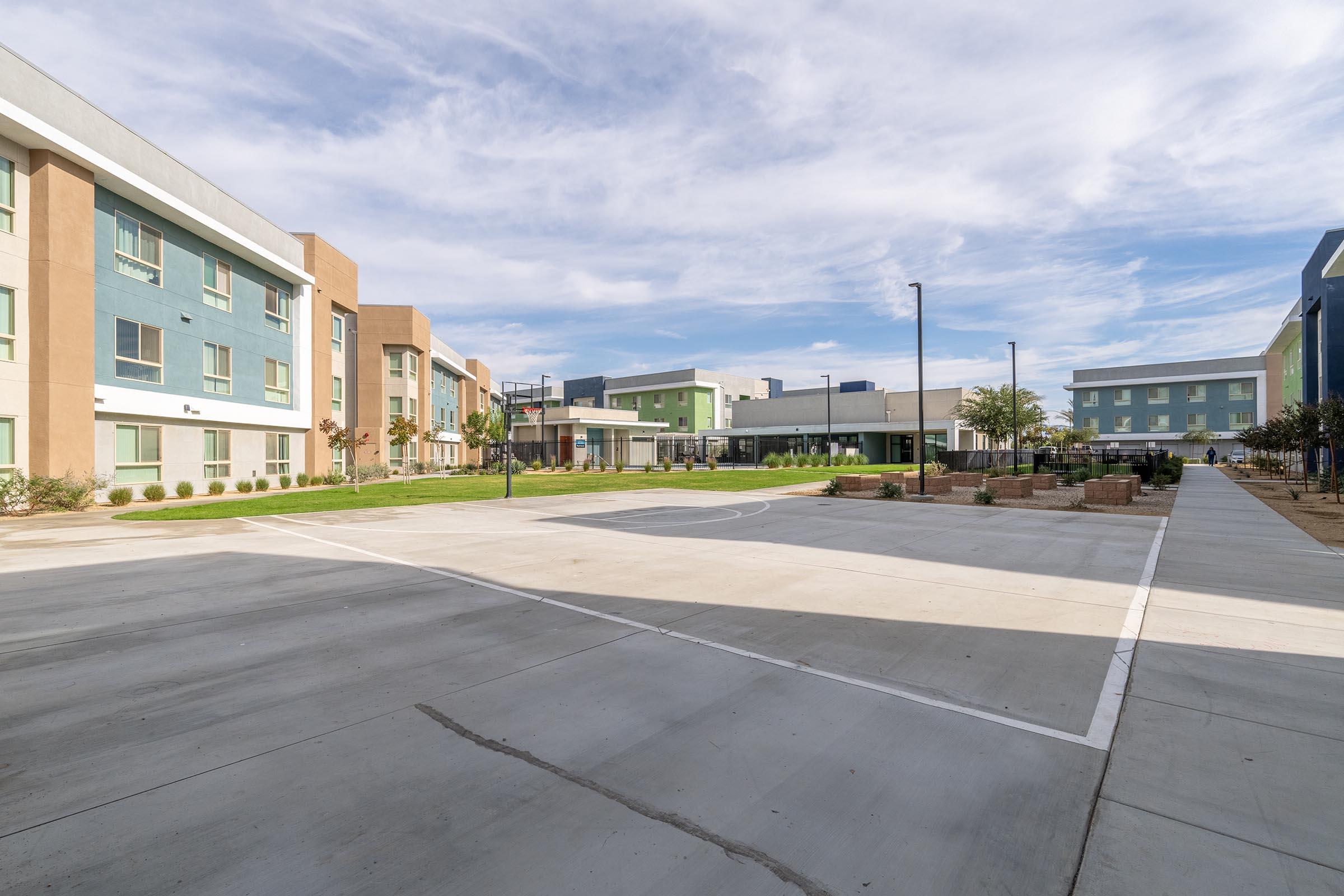 A wide view of a modern educational or residential complex featuring multiple colorful buildings surrounding a paved courtyard. There is a basketball court marked on the pavement, and well-maintained green spaces with shrubs. The sky is partly cloudy, creating a bright and open atmosphere.