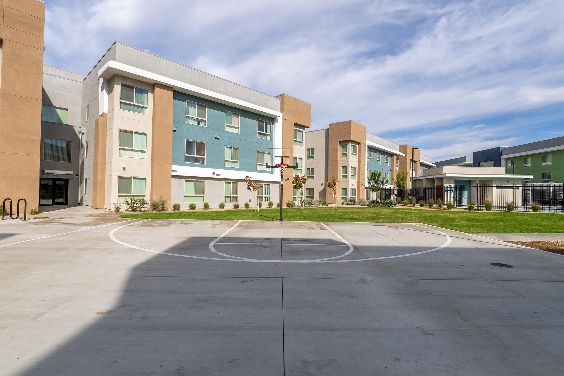 An outdoor basketball court in front of a modern multi-story residential building with a green and beige facade. The court is marked with a hoop and has a smooth concrete surface, surrounded by well-maintained grass and landscaping. The scene is bright with a partly cloudy sky.