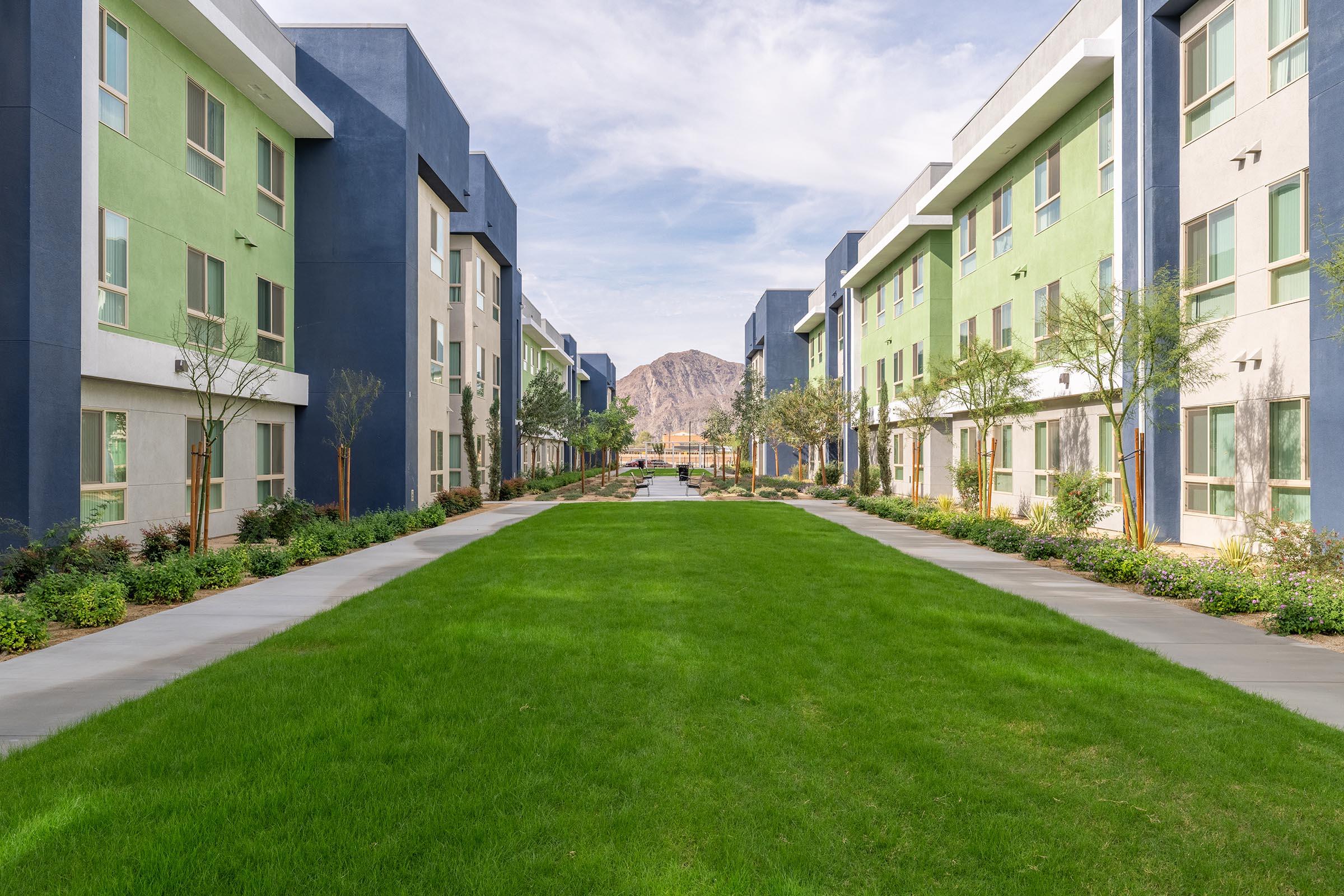A modern residential complex featuring two rows of multi-story buildings in green and blue, framed by manicured lawns and small trees. A pathway leads through the center, with a scenic mountain view in the background under a clear sky.