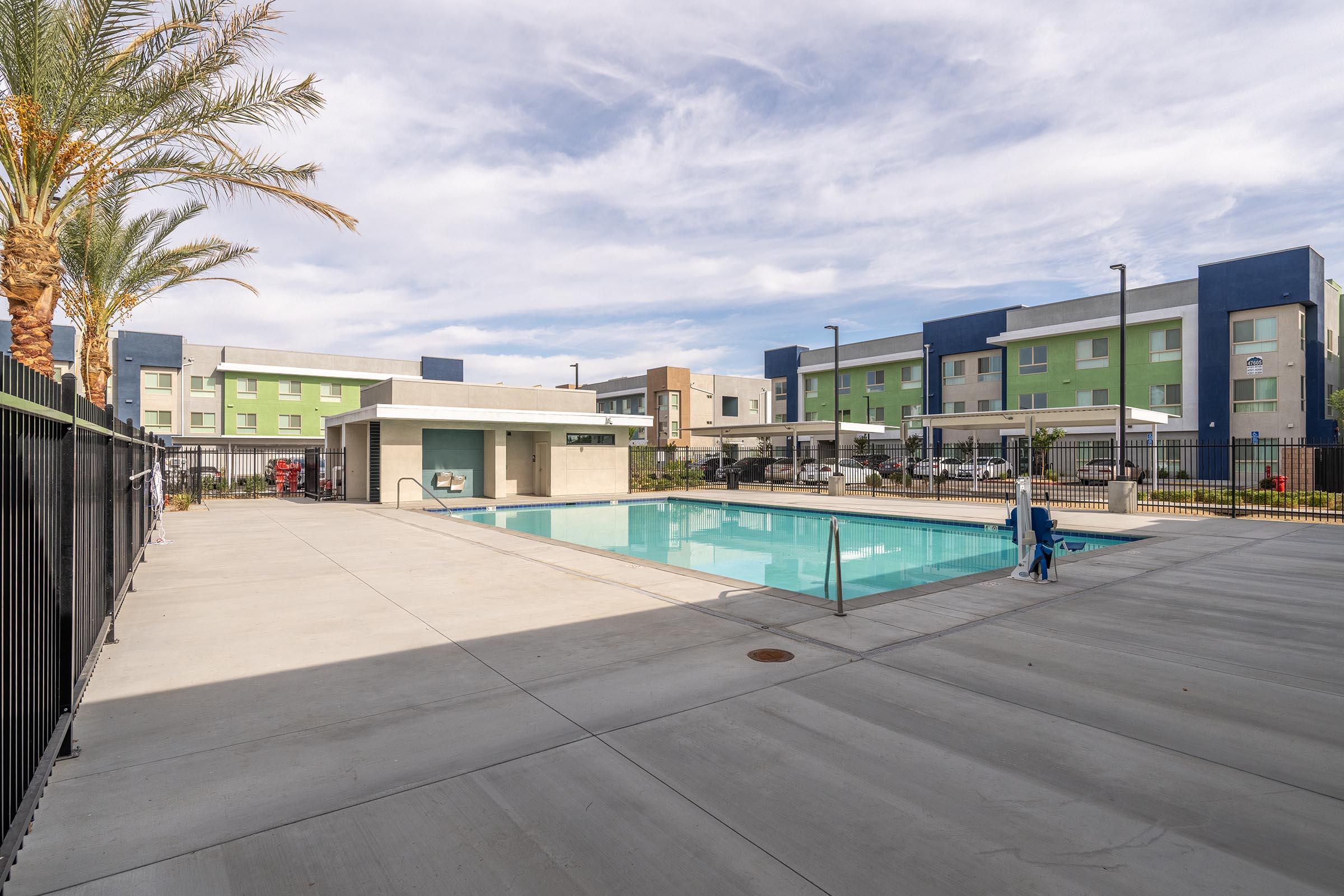 A modern apartment complex featuring a swimming pool area with lounge chairs, surrounded by palm trees. The pool is framed by concrete paving, with additional seating areas visible. The buildings in the background are painted in green and blue, contributing to a contemporary aesthetic.