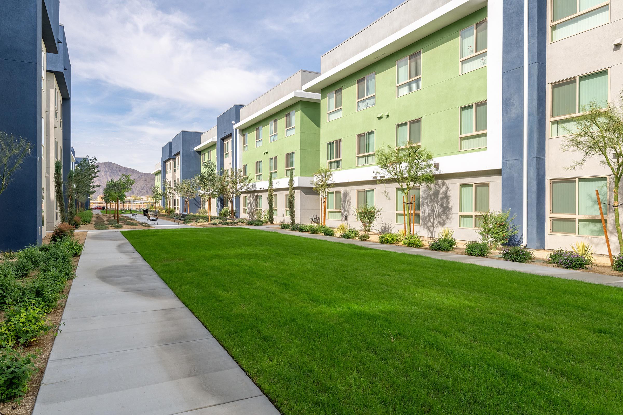 A modern residential building with green and blue exterior walls, featuring multiple floors. The foreground has a neatly manicured lawn with shrubs, trees, and a paved pathway leading through the courtyard, with mountains visible in the background under a partly cloudy sky.
