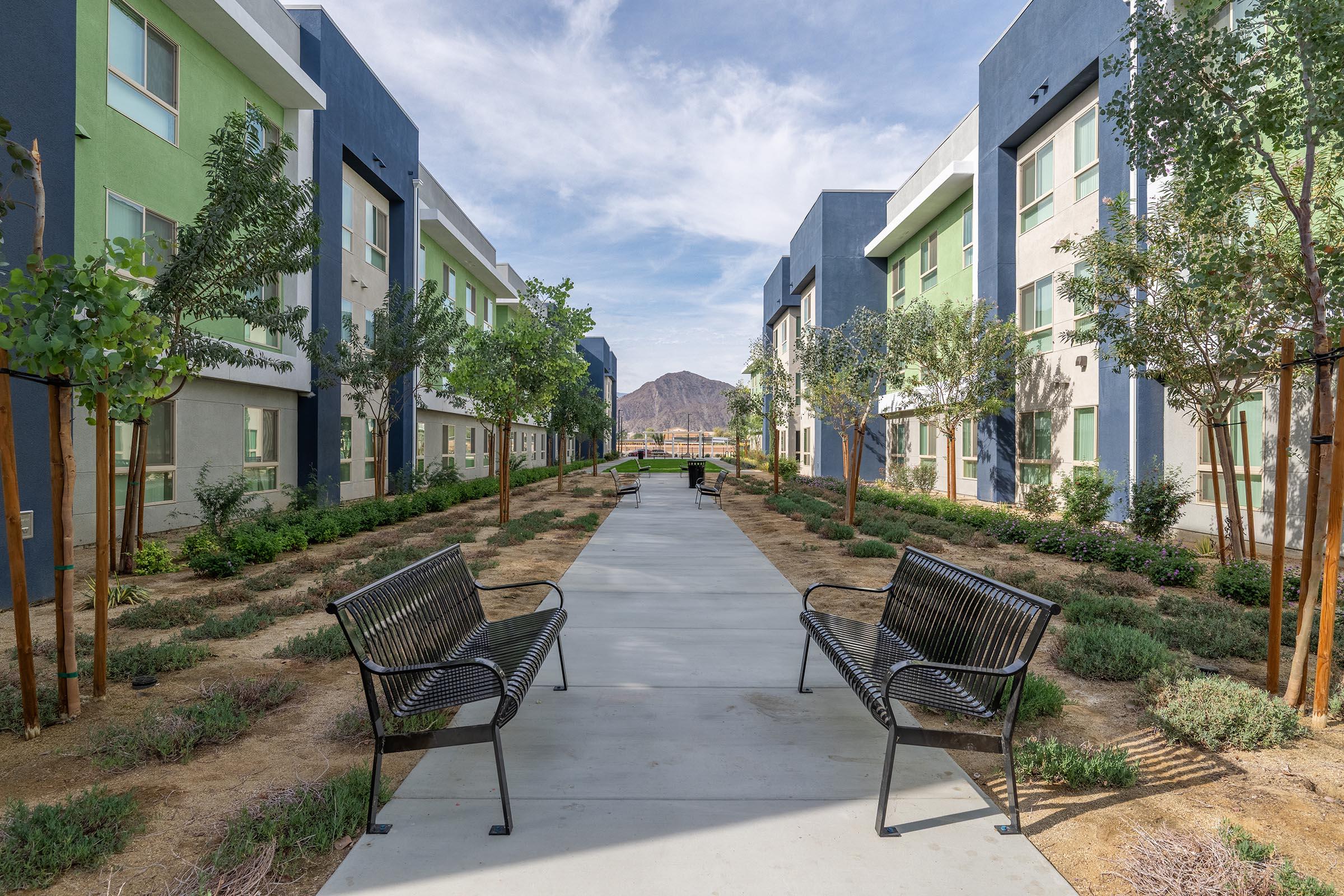 A pathway flanked by benches leads through a landscaped courtyard with greenery and trees, surrounded by modern multi-story buildings painted in shades of green and blue, with a mountain visible in the background under a clear sky.