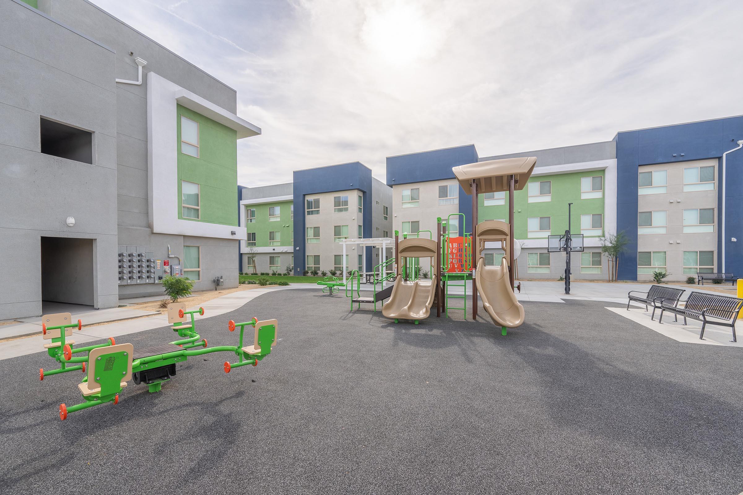 View of a modern playground area featuring two slides and various climbing structures, surrounded by green and gray residential buildings. The ground is covered with a soft surface for safety, and benches are placed nearby for seating. The sky is bright with a few clouds.