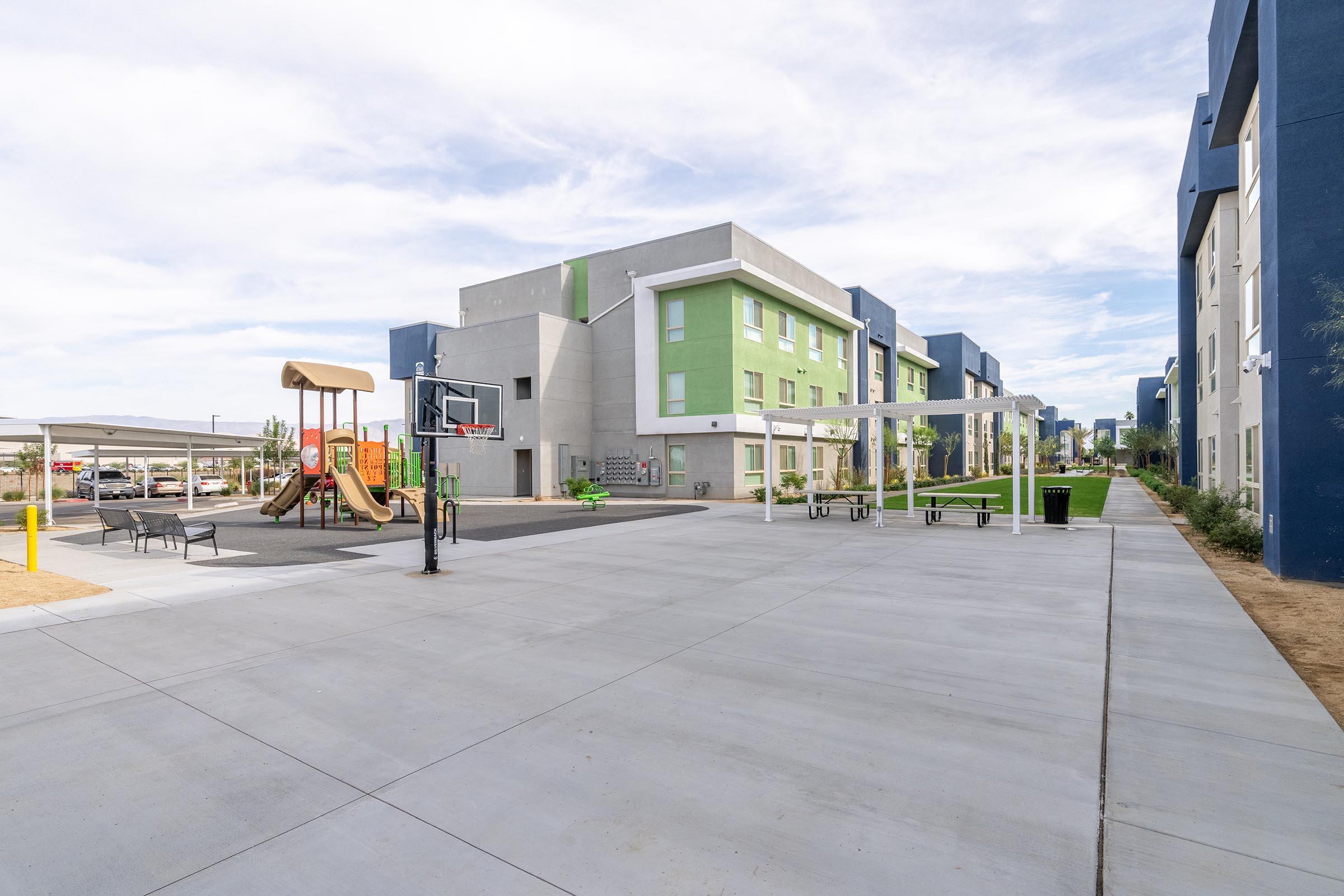 A spacious outdoor area featuring a playground with slides and swings, a basketball hoop, and shaded seating. Surrounding the area are modern apartment buildings painted in blue and green, with a clear sky overhead. The surface is paved, providing a clean and open environment for recreation and relaxation.