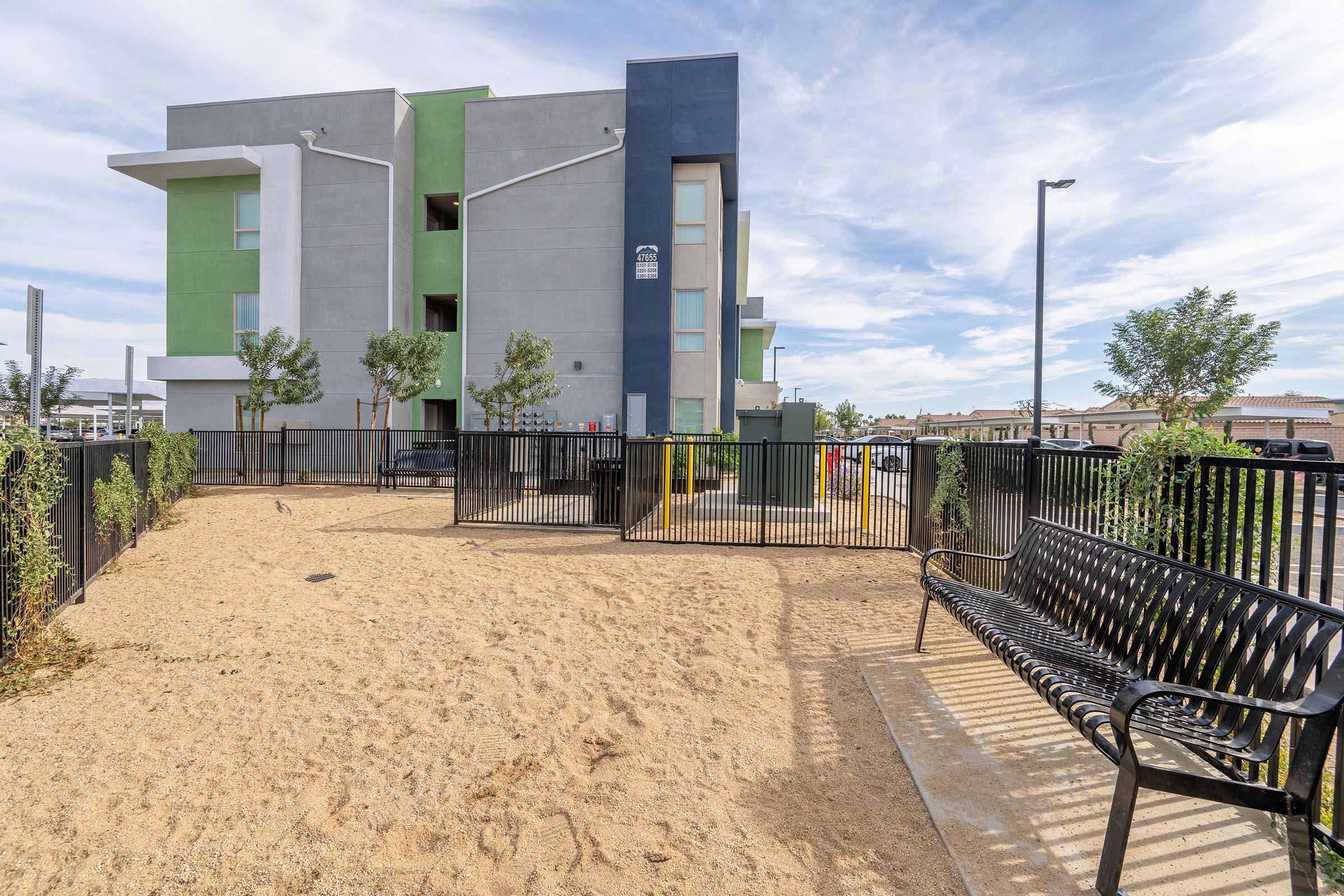 A view of a sandy area with a black bench and small trees, located near a modern multi-story apartment building with a mix of gray and green exterior. The sky is partly cloudy, and a fenced area is visible in the background, suggesting a residential environment.