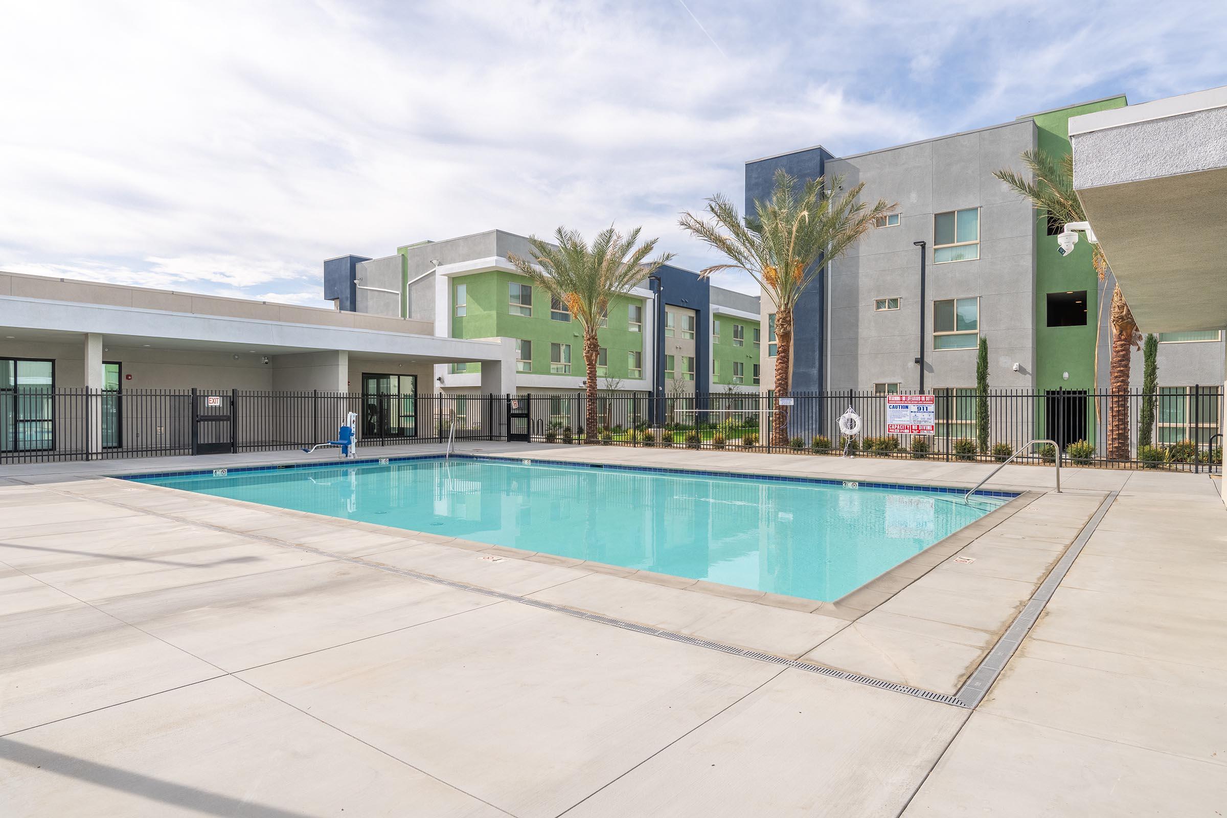 A clean, well-maintained swimming pool surrounded by concrete and palm trees, with modern apartment buildings in the background. The pool area is enclosed by a black fence, and there are lounge chairs arranged around the poolside. A clear sky indicates a bright day.