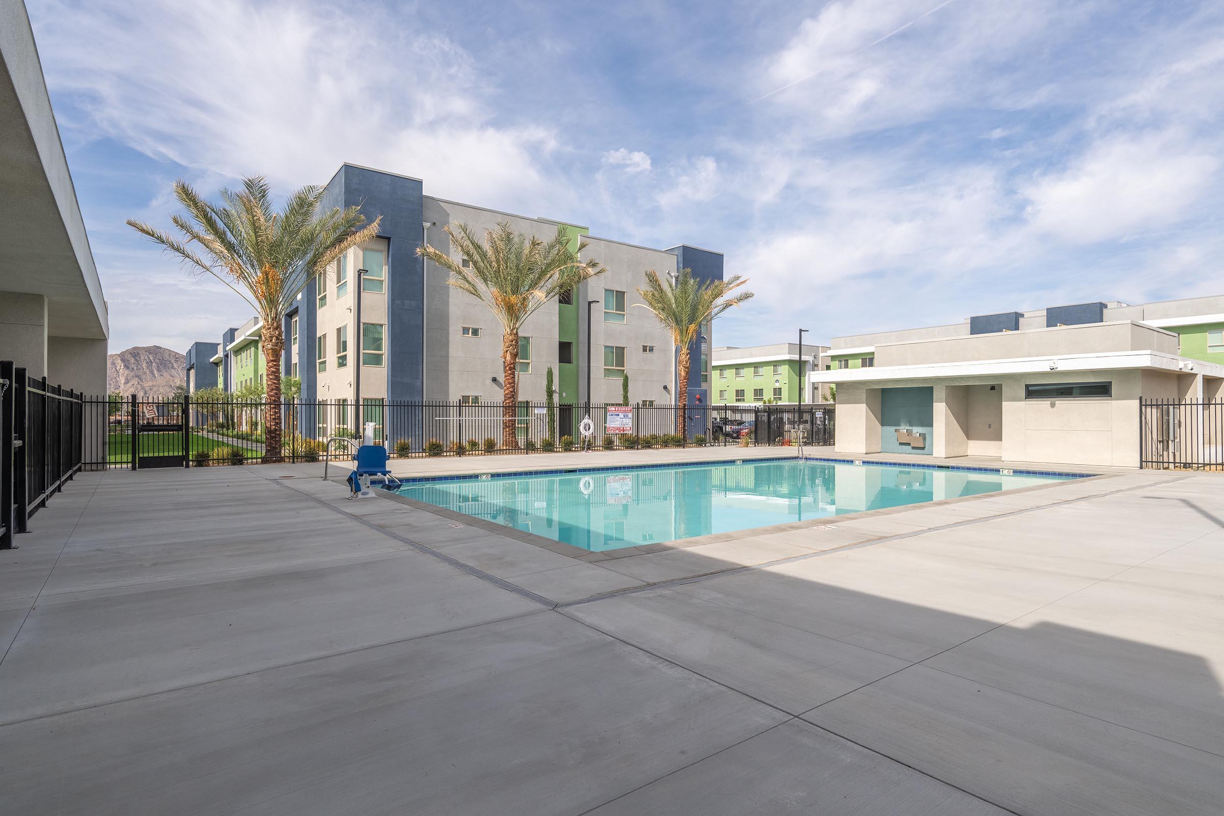 View of a modern apartment complex featuring a clear swimming pool surrounded by a concrete deck. Palm trees are planted nearby, and the buildings have a mix of gray and green exteriors. The sky is partly cloudy, creating a bright and inviting atmosphere.