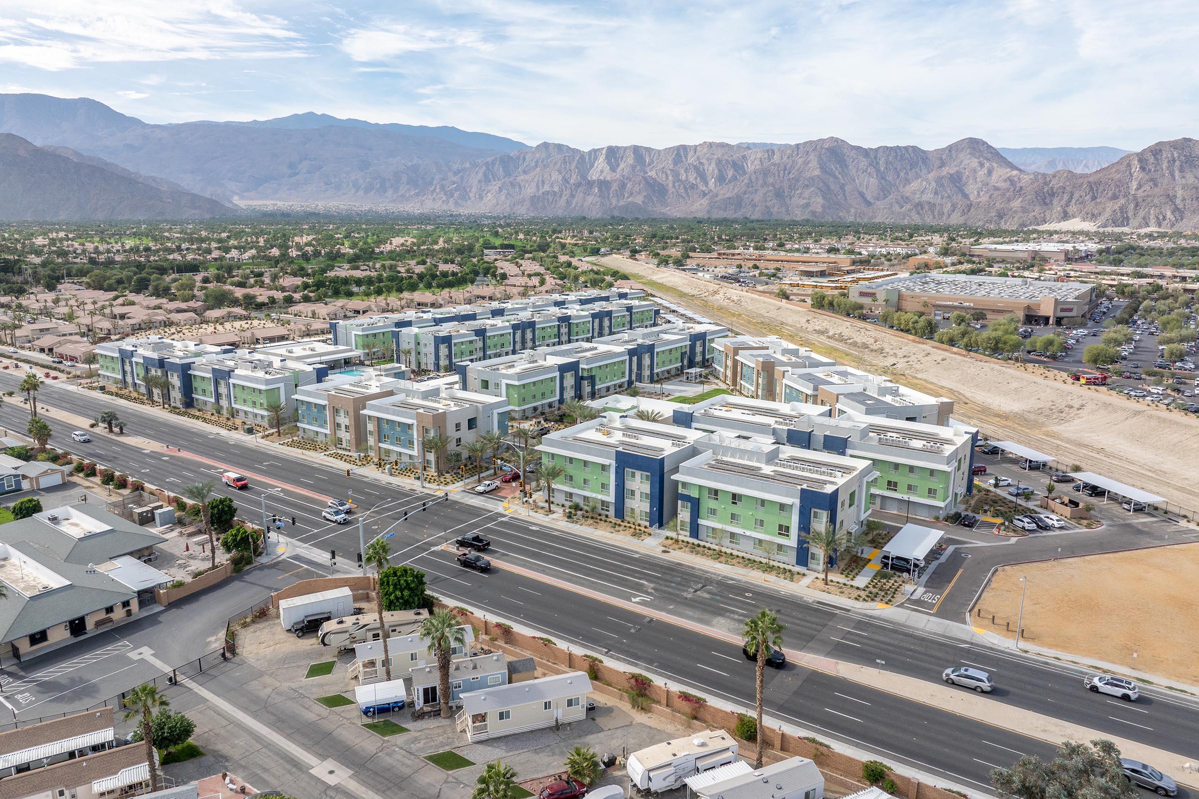 Aerial view of a modern multi-story residential complex set against a mountainous backdrop. The building features a mix of green and blue colors. In the foreground, a busy road runs parallel to the complex, with various vehicles and palm trees visible. Sparse vegetation and desert terrain are present beyond the buildings.