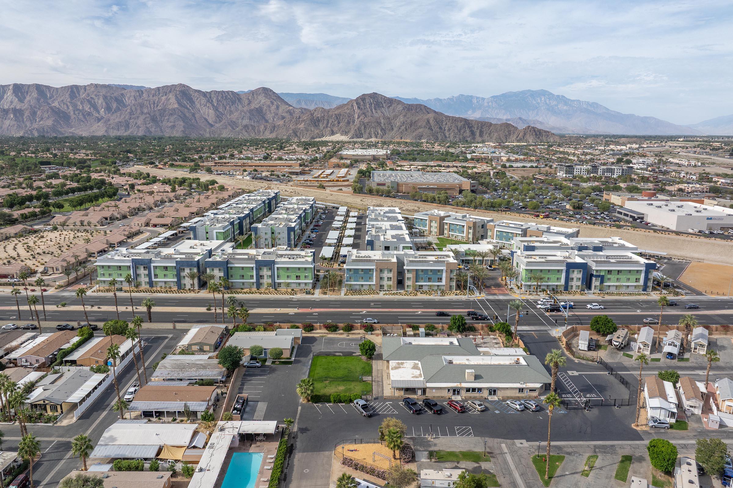 Aerial view of a modern residential complex with multiple buildings, surrounded by mountains and open land. In the foreground, there are parking lots and some trees, while the background features a valley with distant mountains under a partly cloudy sky.