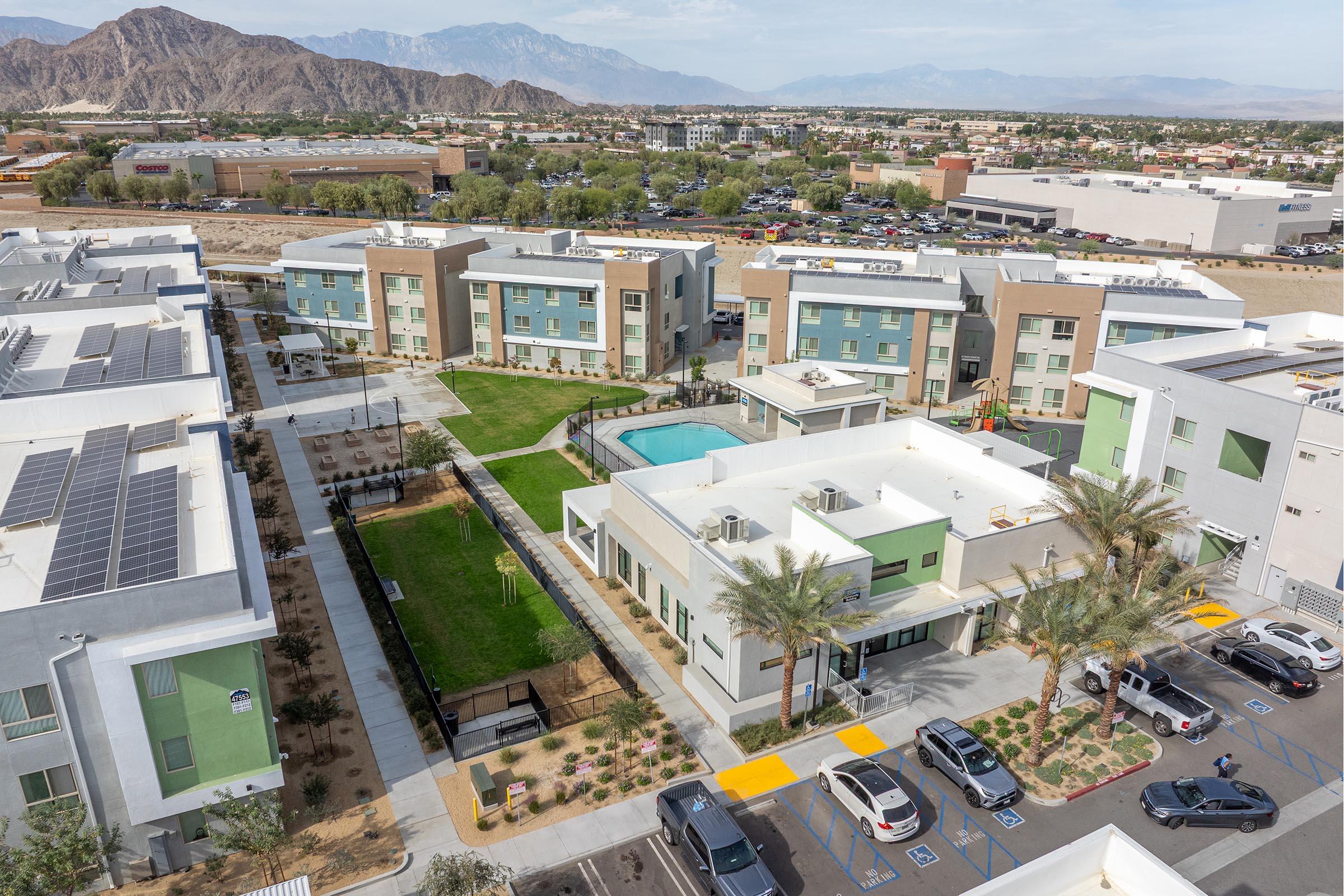Aerial view of a modern apartment complex featuring several multi-story buildings with green and beige facades, a landscaped courtyard with a swimming pool, and surrounding parking lots. Background shows mountains and a distant urban area with various buildings.