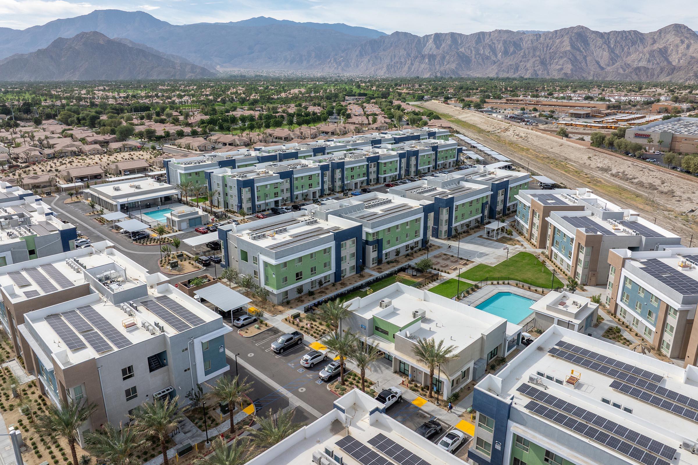 Aerial view of a modern apartment complex featuring multiple buildings, solar panels on rooftops, landscaped areas, swimming pools, and surrounding mountains. In the foreground, palm trees line the pathways, and various residential and commercial structures are visible nearby.