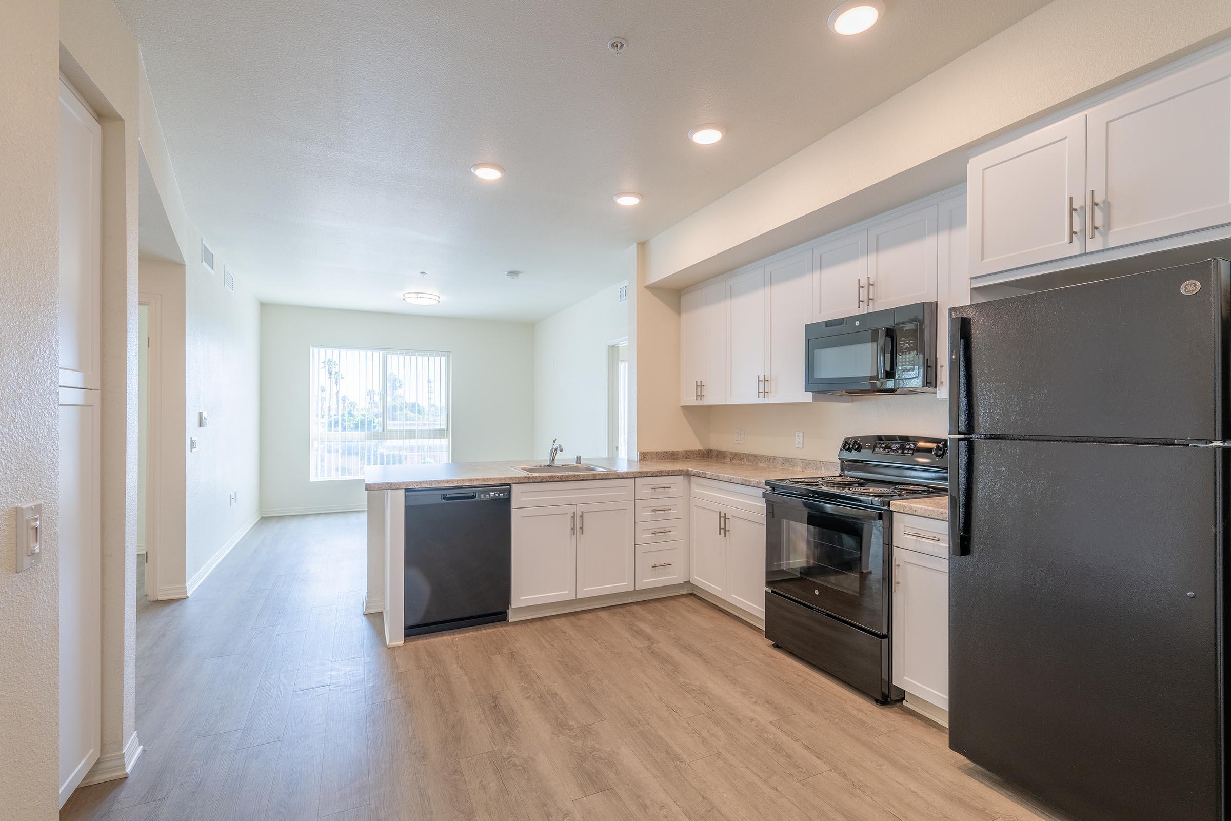 Modern kitchen interior featuring white cabinetry, stainless steel appliances including a microwave and dishwasher, and a black refrigerator. The space has a light-colored countertop and laminate flooring, illuminated by recessed lighting. A large window offers natural light, with an open layout leading to a living area.