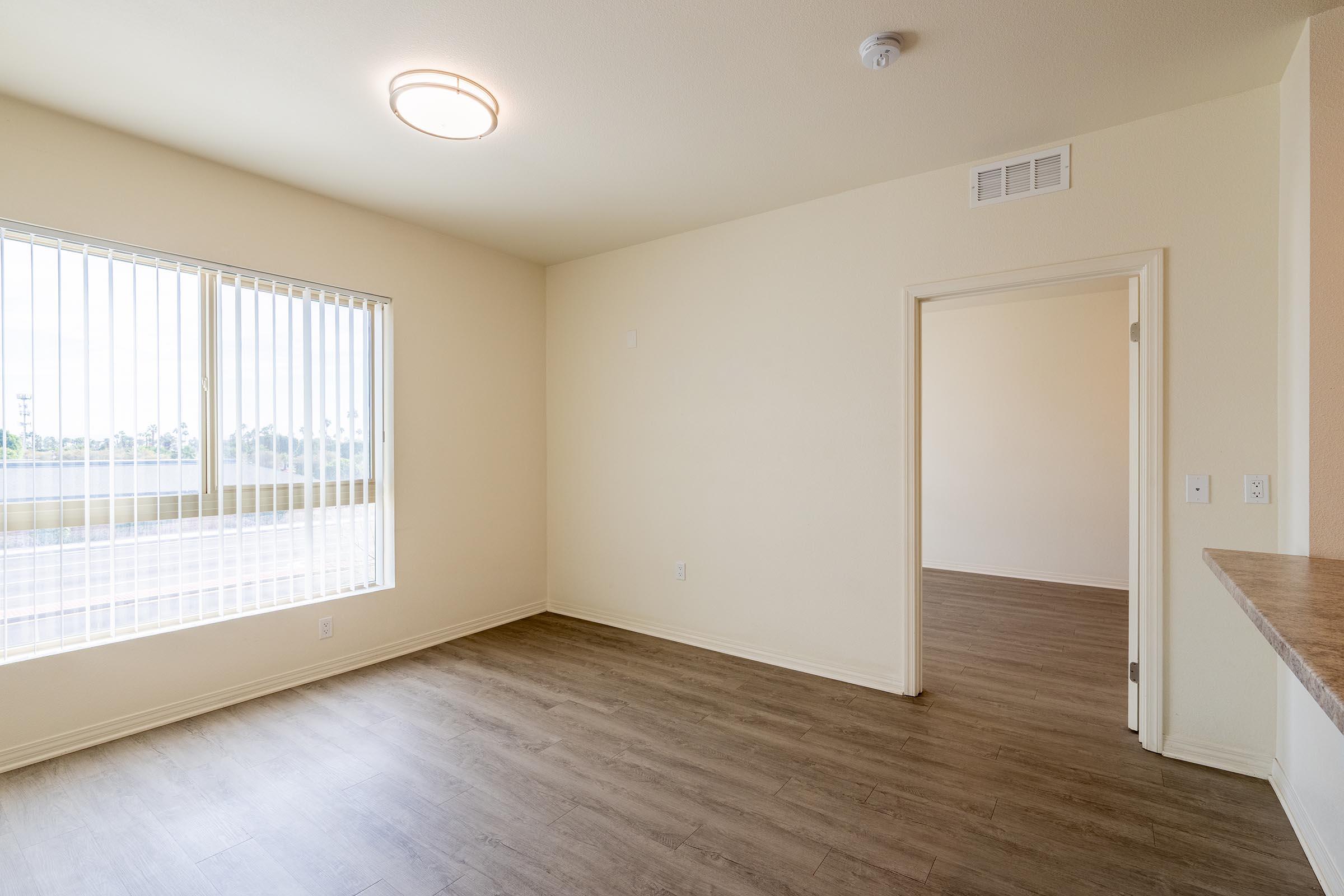 A spacious, empty room with light-colored walls and modern wood-look flooring. A large window with vertical blinds lets in natural light, and a doorway leads to another room. The ceiling features a simple round light fixture, and there is a small area of counter space visible.