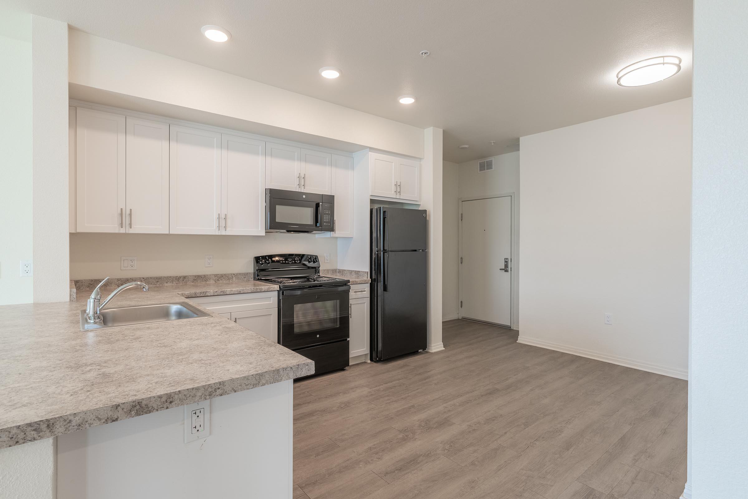 A modern kitchen interior featuring white cabinets, a stainless steel microwave, a black refrigerator, and a black stove. The countertop has a light marble finish. There are overhead lights and a partially visible entrance door in the background. The flooring is light wood laminate.